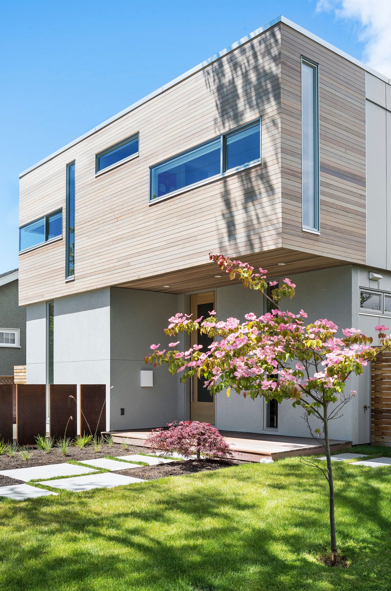 Modern two-story home with horizontal wood siding, large windows, concrete path, and blooming pink dogwood tree.