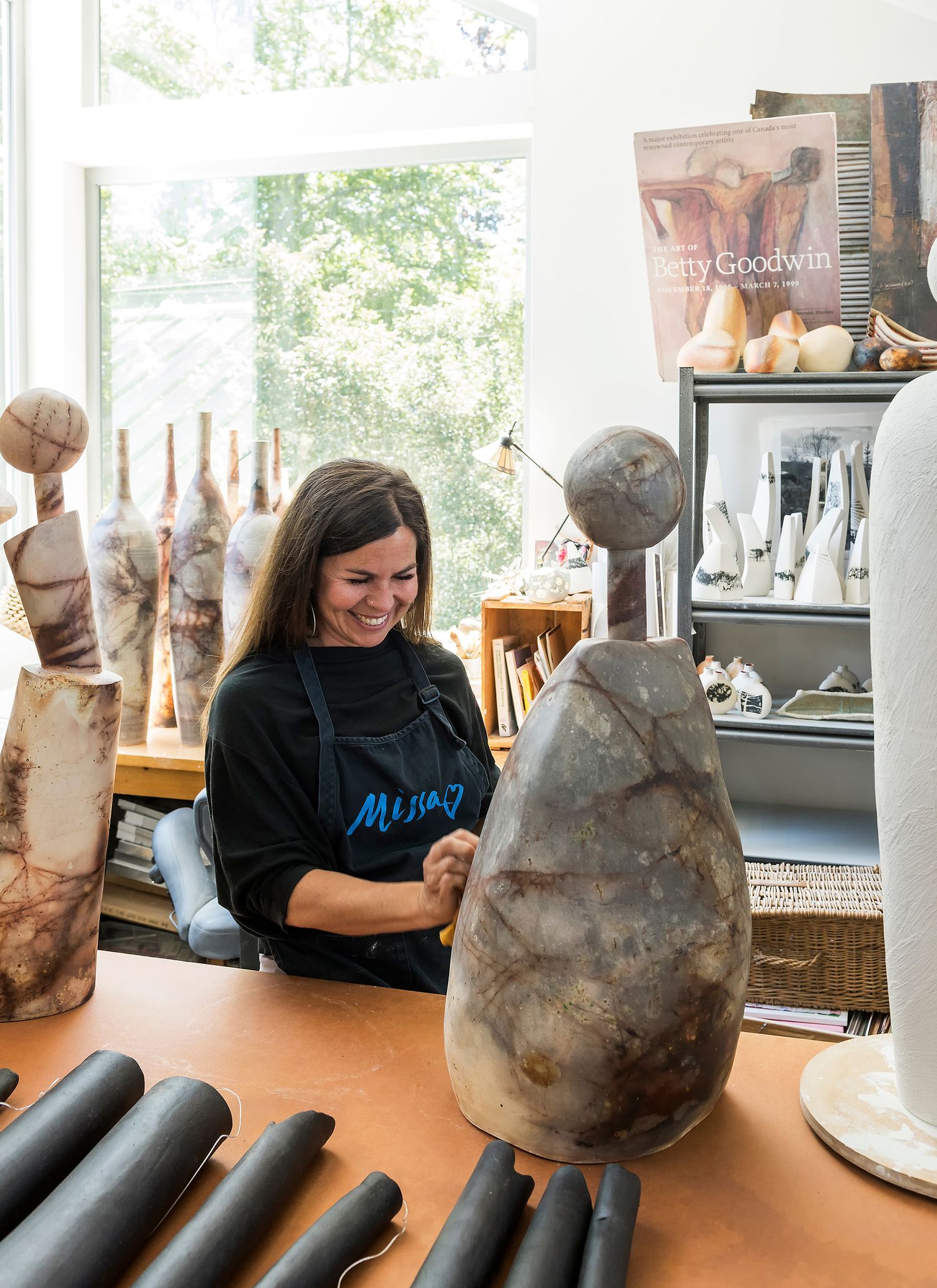 Smiling woman in black apron working on ceramic sculptures in bright art studio workshop.