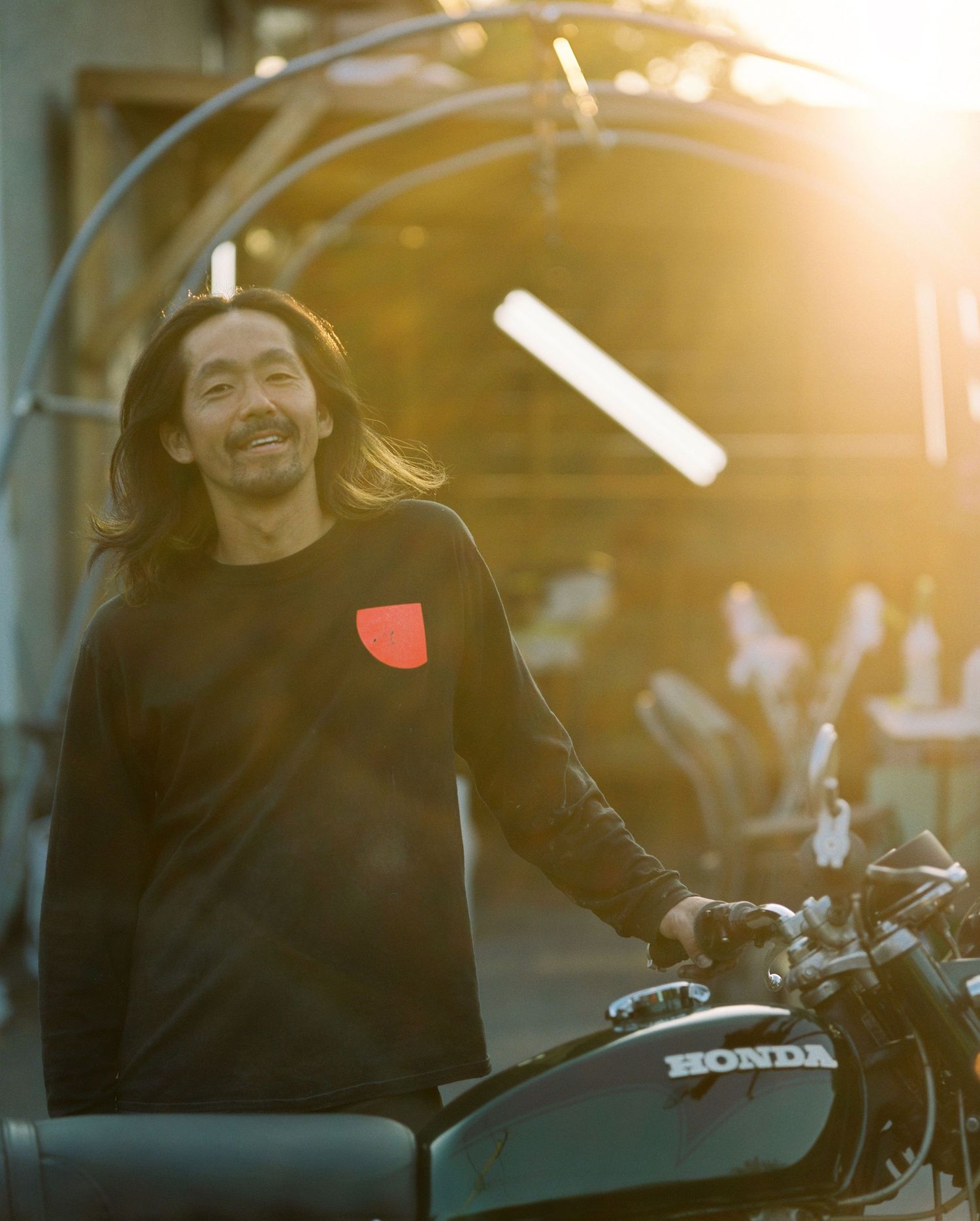 Long-haired man smiles beside a Honda motorcycle in warm golden sunset light.