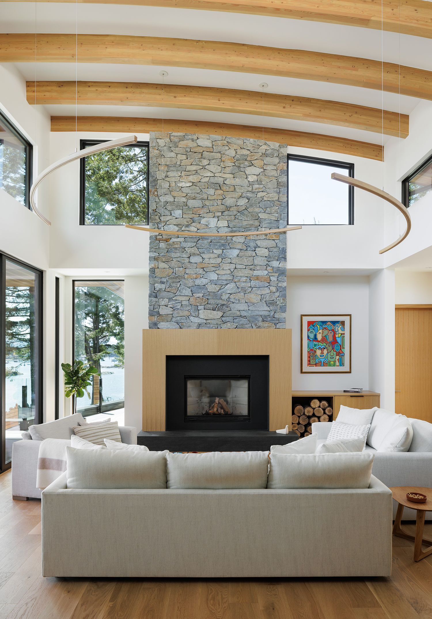 Bright contemporary living room featuring stone fireplace, curved ceiling beams, white sectional sofa, and large windows.