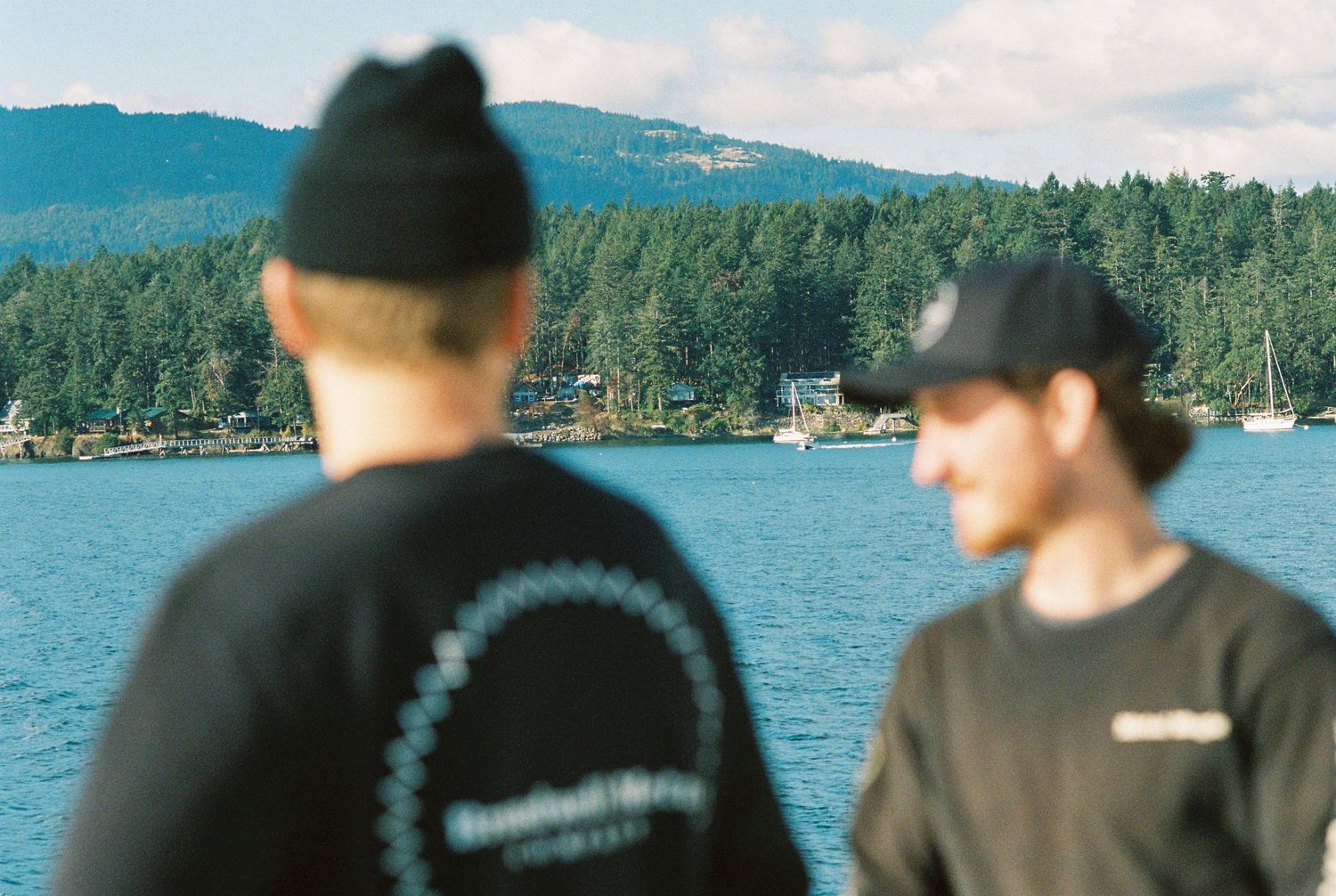 Two people in black sweatshirts stand on a ferry deck overlooking a forested coastal inlet.