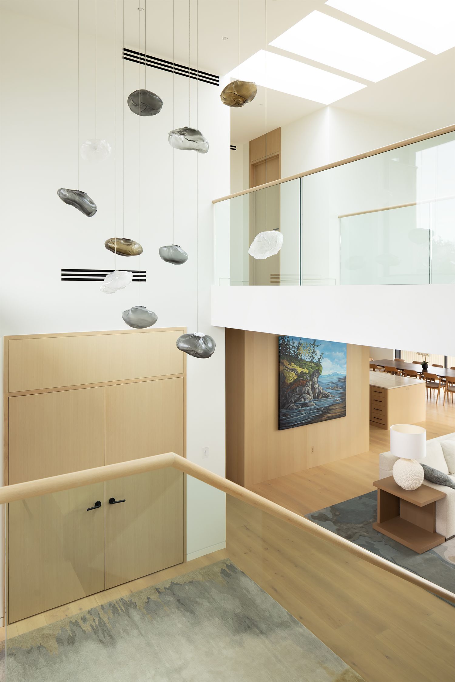 Modern two-story interior with stone mobile sculpture, light wood cabinetry, glass railing, and skylight above staircase.