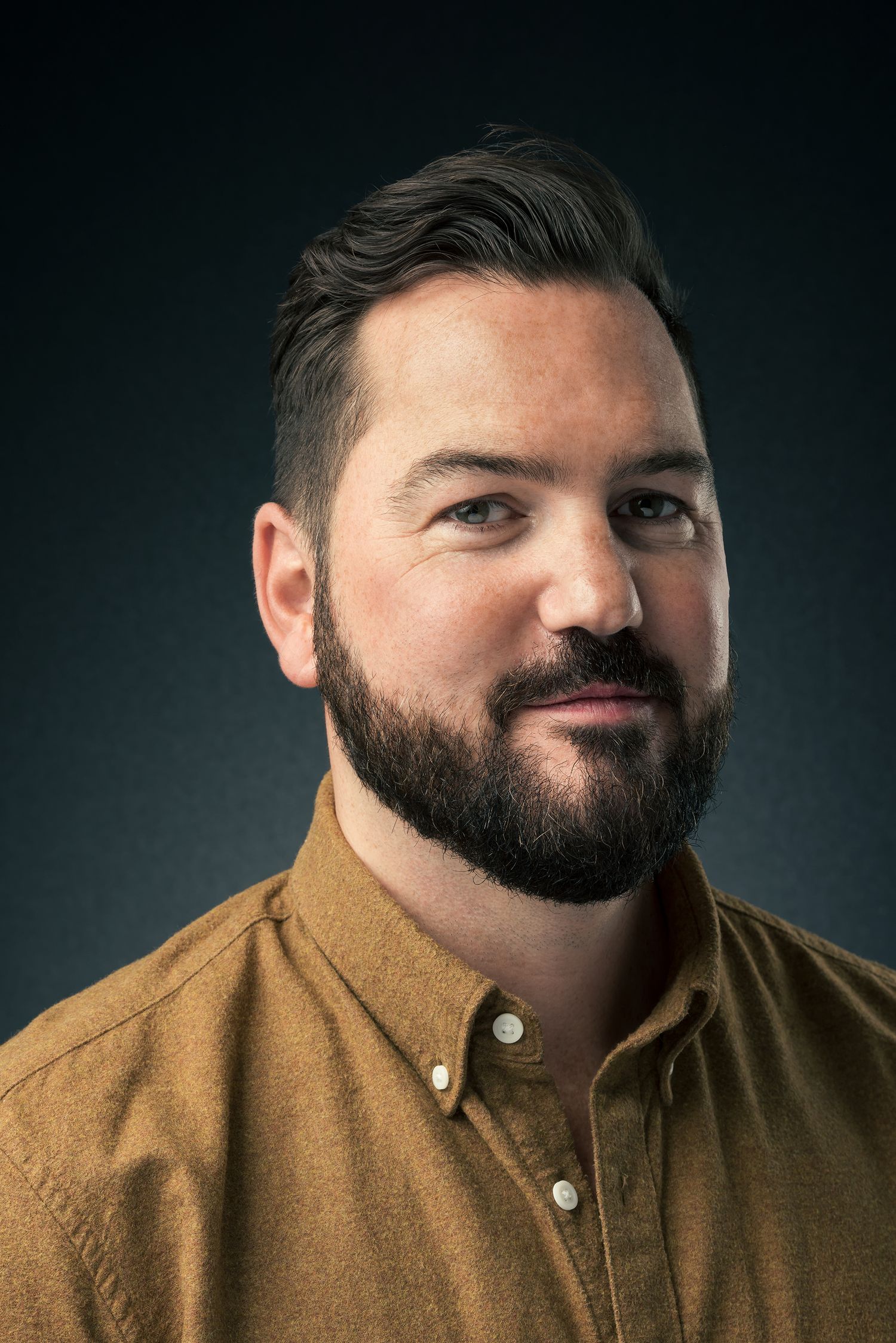 Bearded man in mustard shirt poses for professional headshot against dark background.