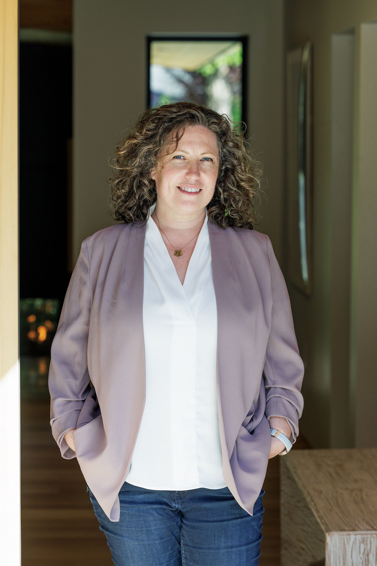 Smiling woman with curly hair wearing lavender blazer and white blouse posing in a modern interior space.
