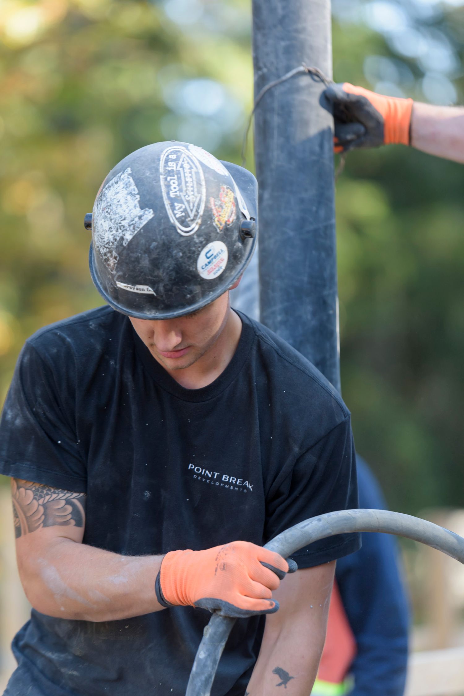 Tattooed construction worker in black hard hat guides a concrete pump hose at a building site.