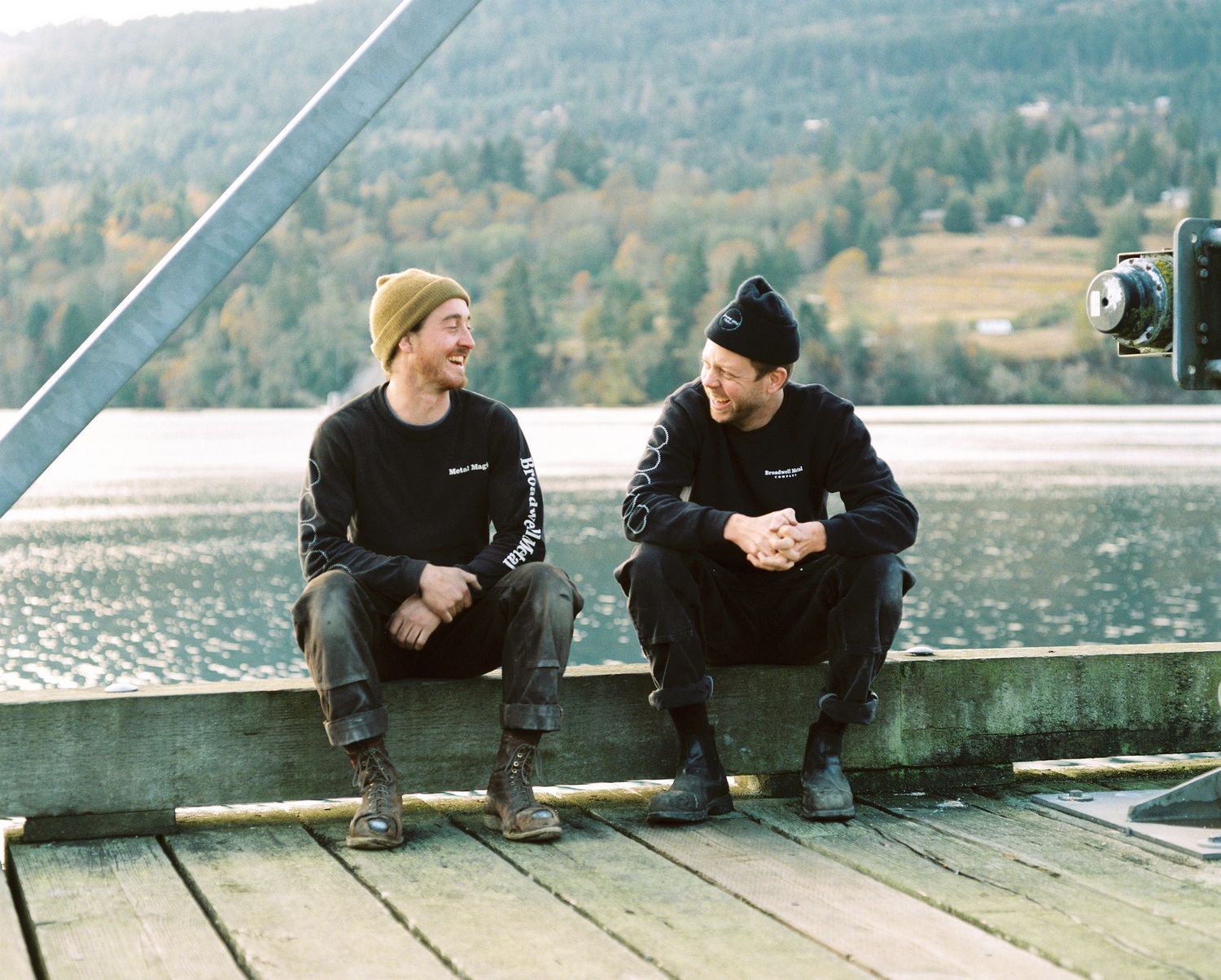 Two men in black beanies and sweatshirts sit laughing on a wooden dock with mountain backdrop.