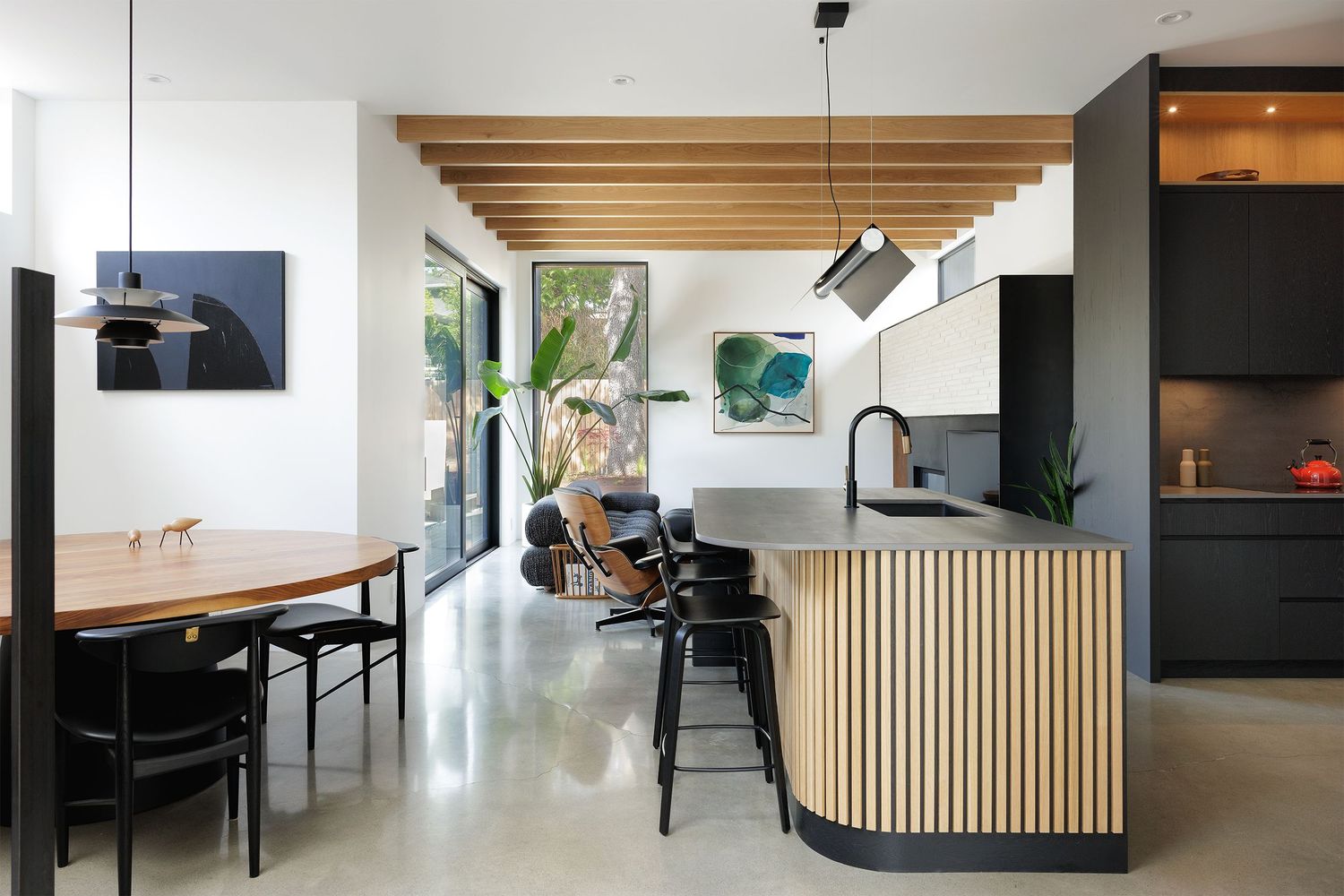 Modern open-plan kitchen and dining with wood slat island, exposed beam ceiling, polished concrete floors, and black accents.