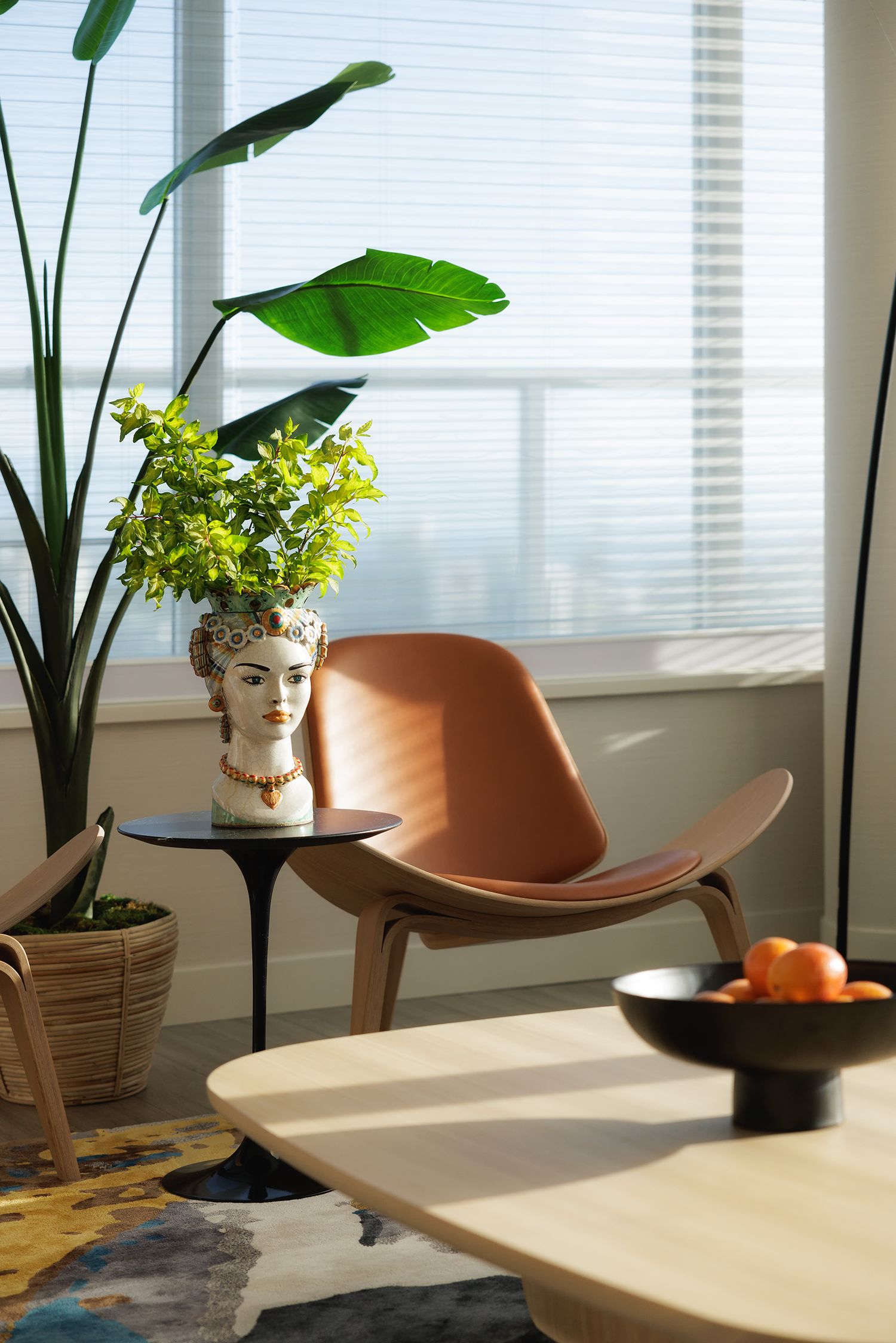 Modern living room corner with leather shell chair, face planter, tropical plants, and natural light through window blinds.