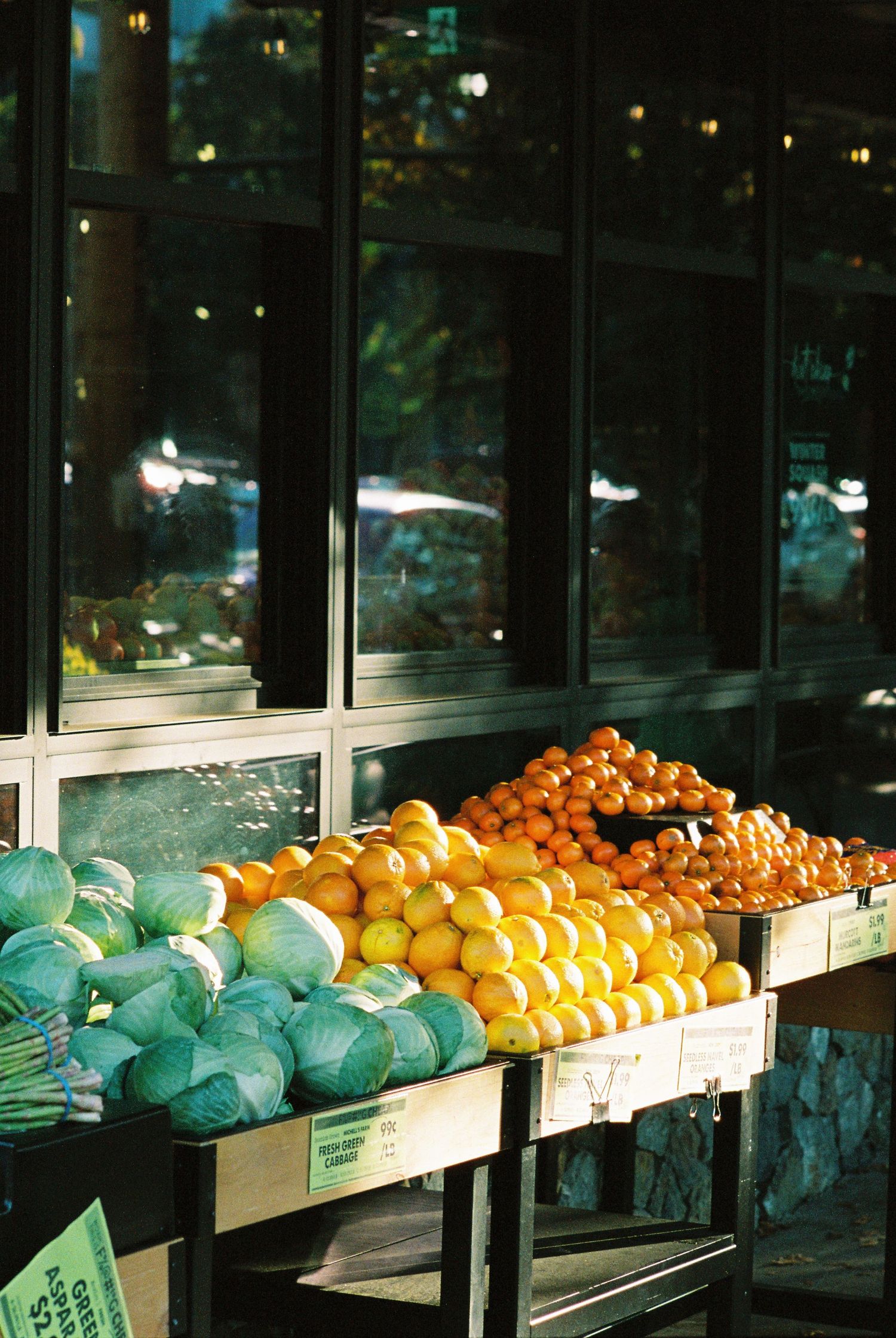 Outdoor produce stand displaying cabbage, lemons, and oranges in sunlight outside a dark glass storefront.