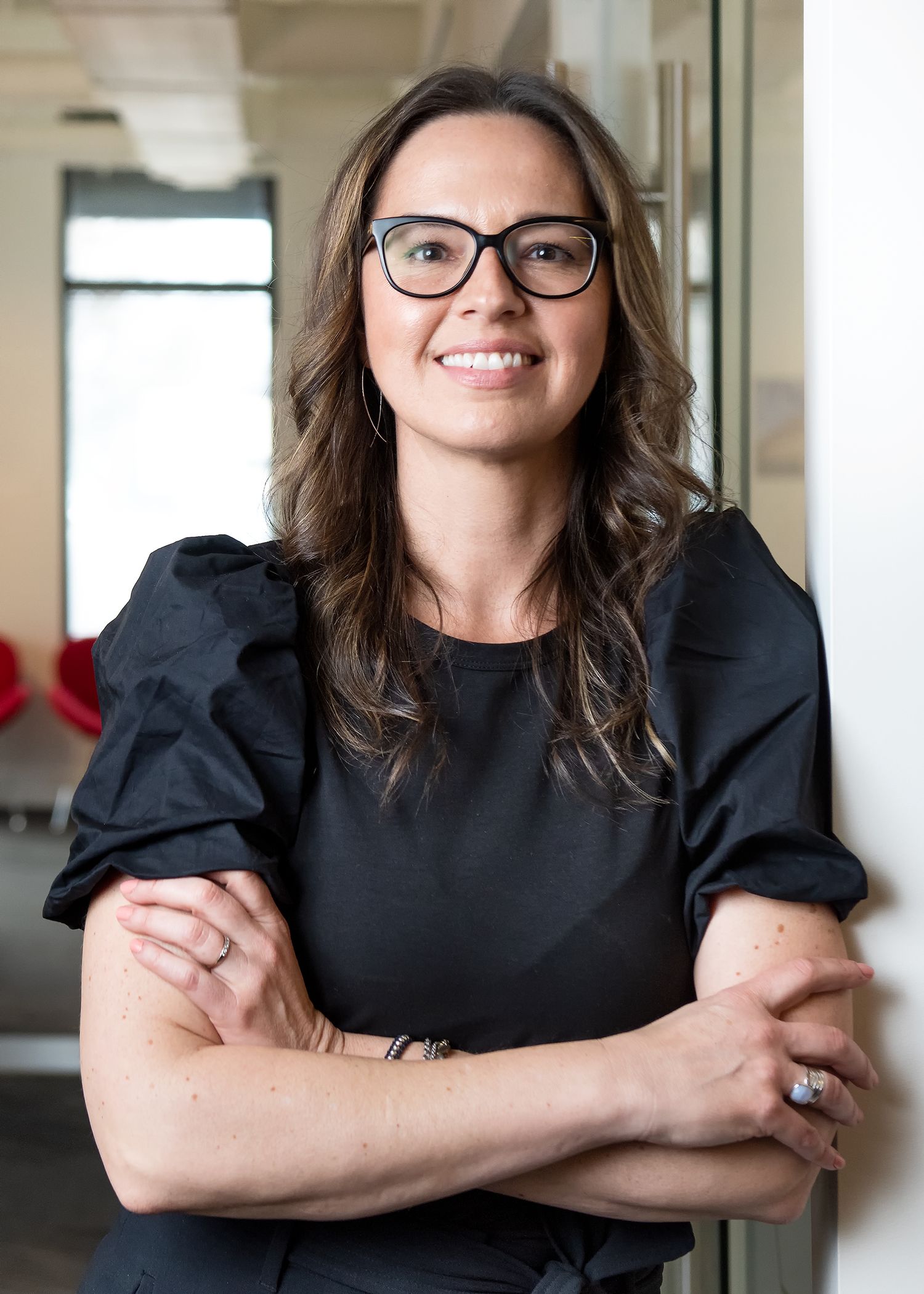 Smiling brunette woman in black top with glasses leans against wall in bright office.