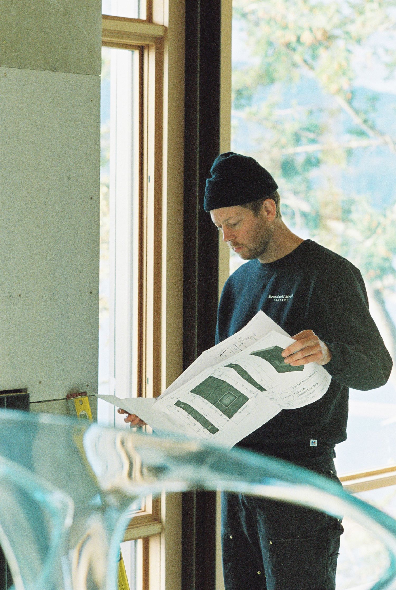 Man in black beanie reviewing architectural blueprints by a window in a construction site interior.