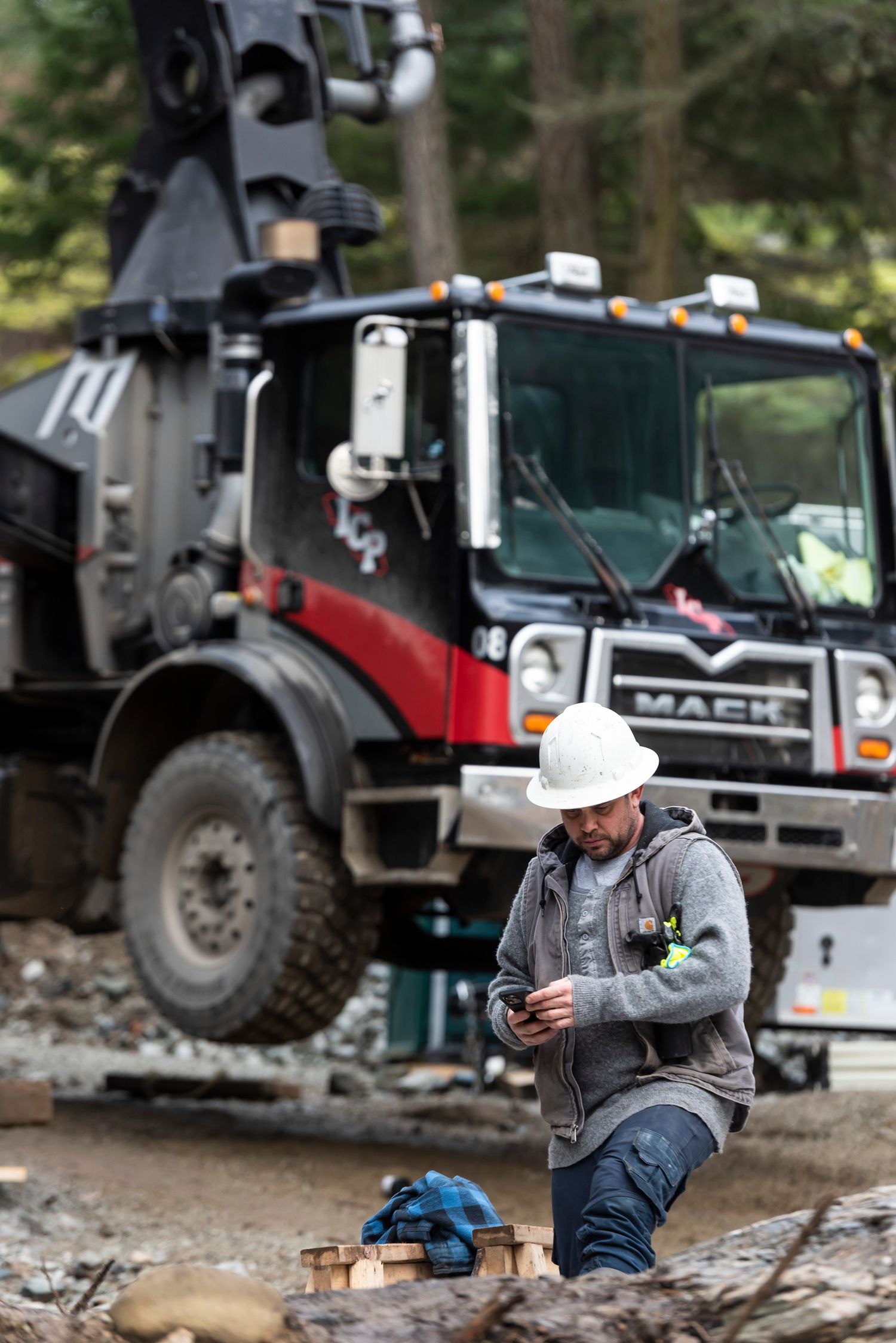 Construction worker in white hard hat kneeling and using a phone in front of a large Mack truck on a job site.