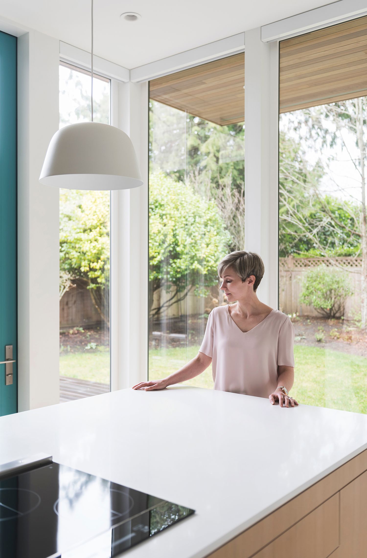 Woman with short hair standing at white kitchen island looking out large window to garden.