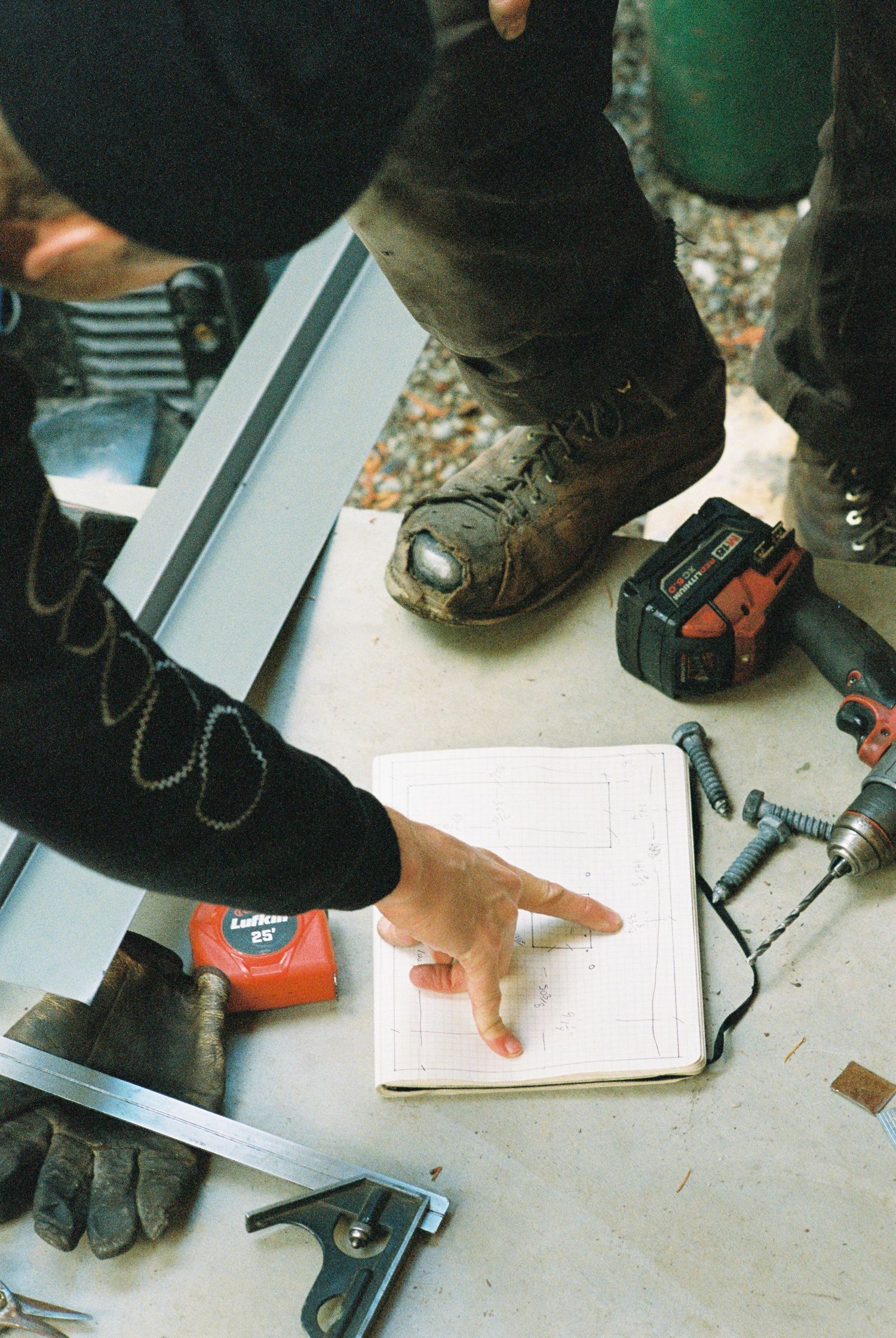 Construction worker pointing at notebook plans surrounded by tools and a tape measure on the ground.
