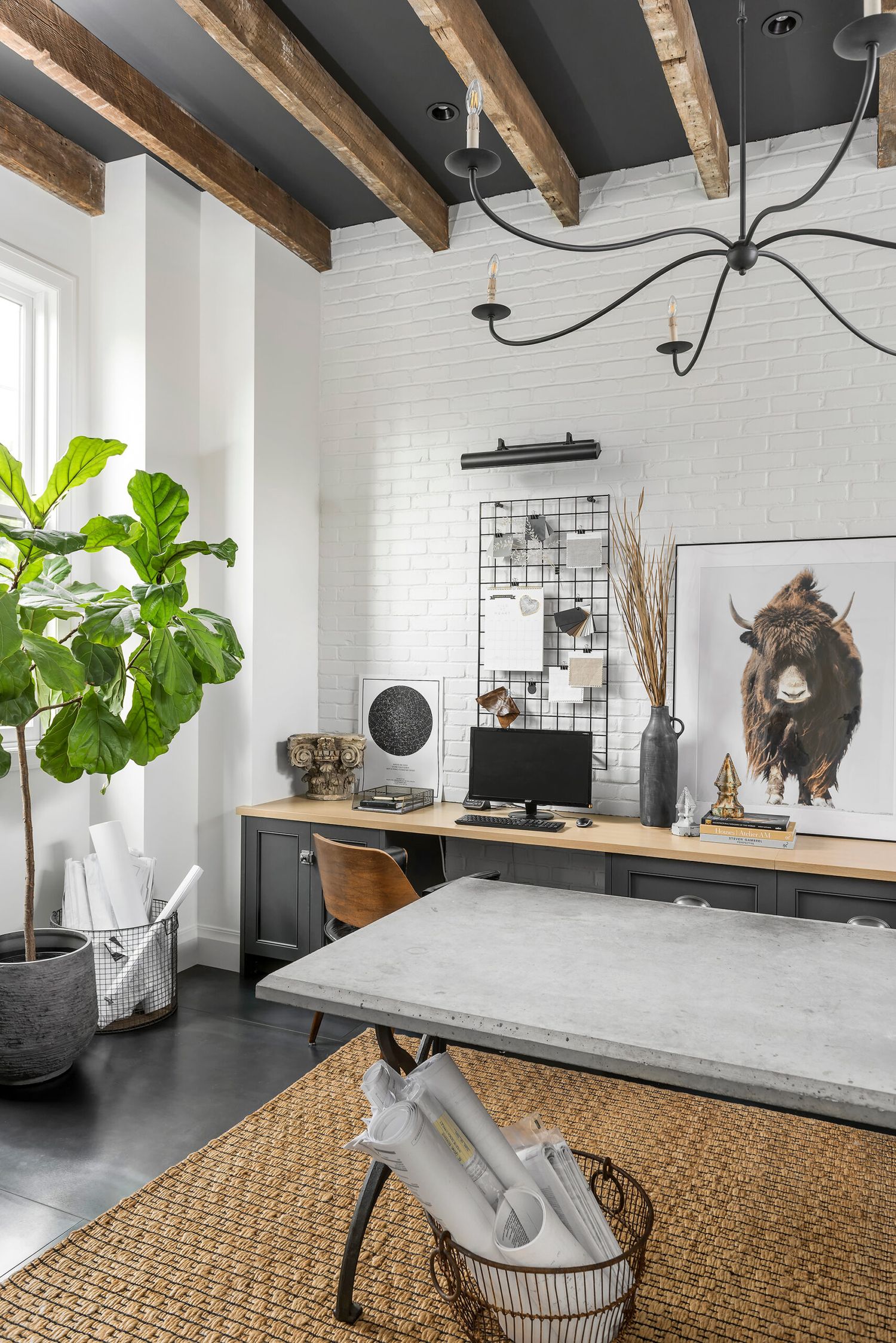 Industrial home office with rustic wood beam ceiling, brick wall, concrete table, dark cabinets, and a fiddle leaf fig tree.
