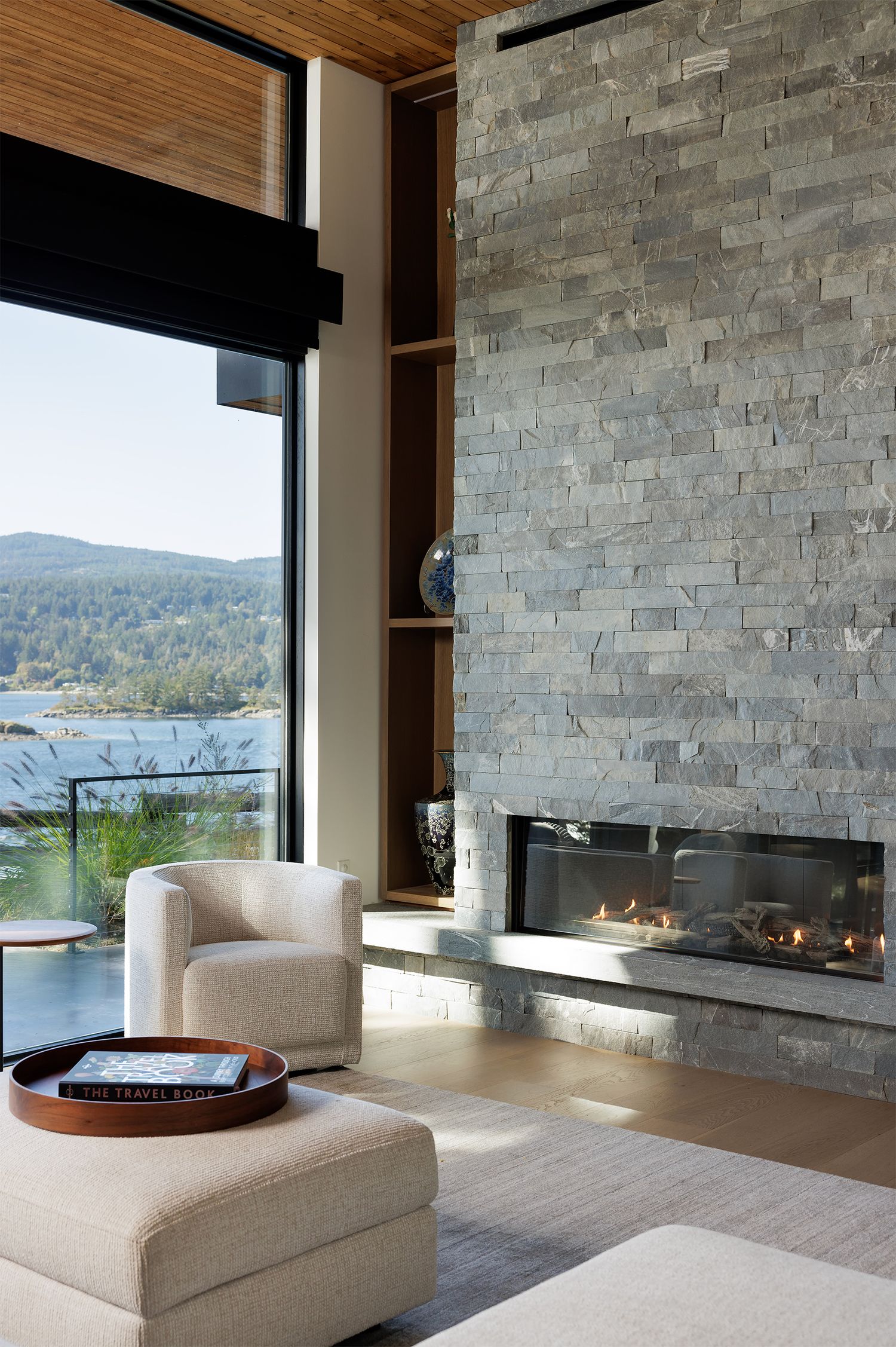 Modern living room with stone fireplace, white armchair, and panoramic mountain lake views through floor-to-ceiling windows.
