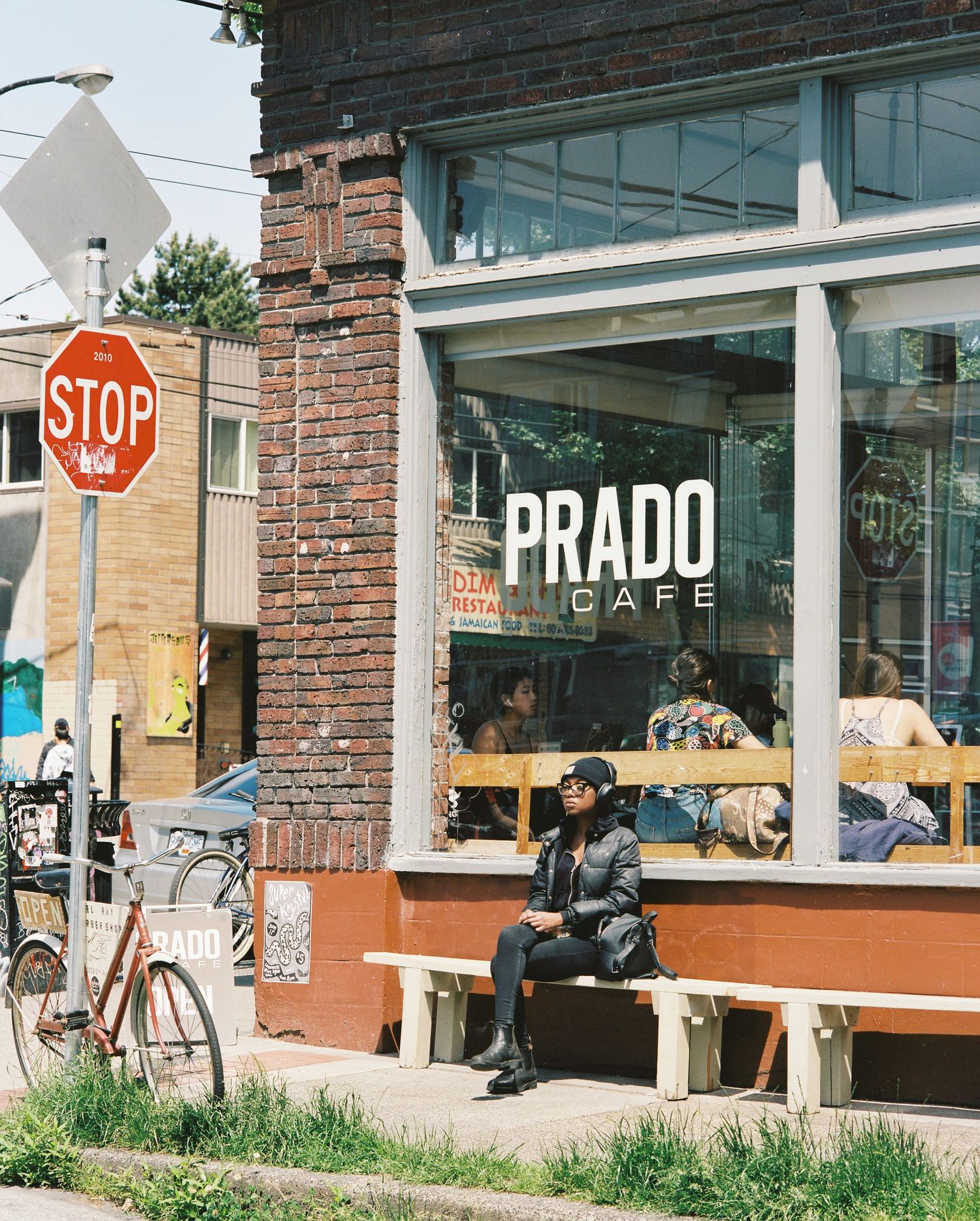 Person sits on bench outside Prado Cafe corner shop on sunny urban street.