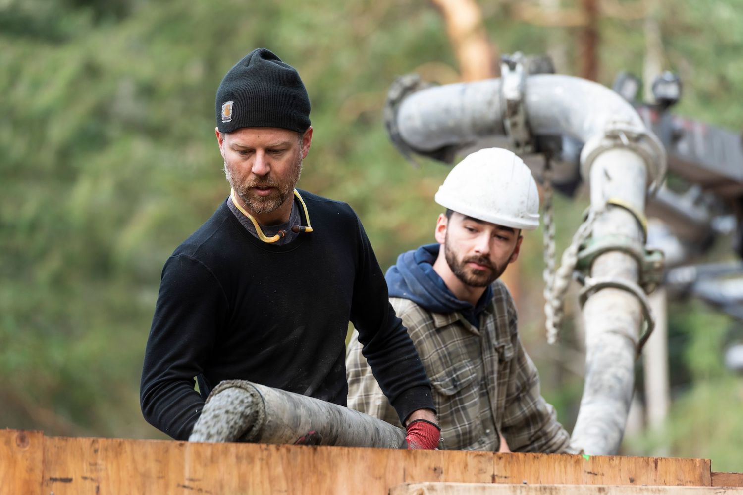 Two male construction workers manage a concrete pump hose on a job site with pine trees in the background.