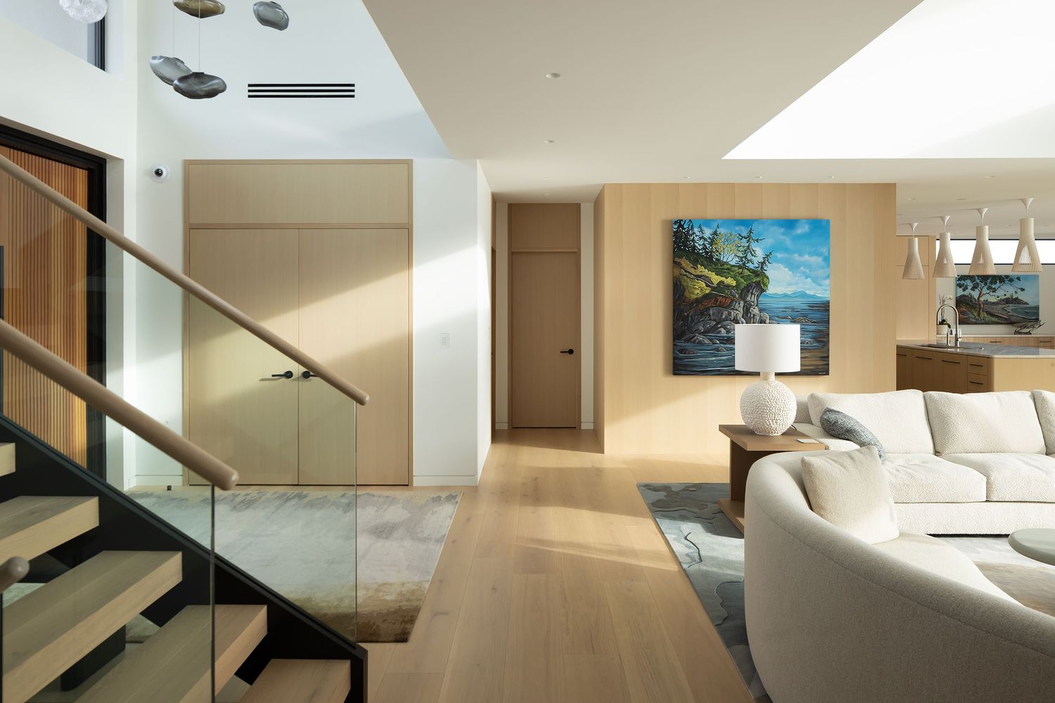 Modern open-plan living room with glass stair railing, light wood floors, white sofa, and vibrant blue landscape painting.