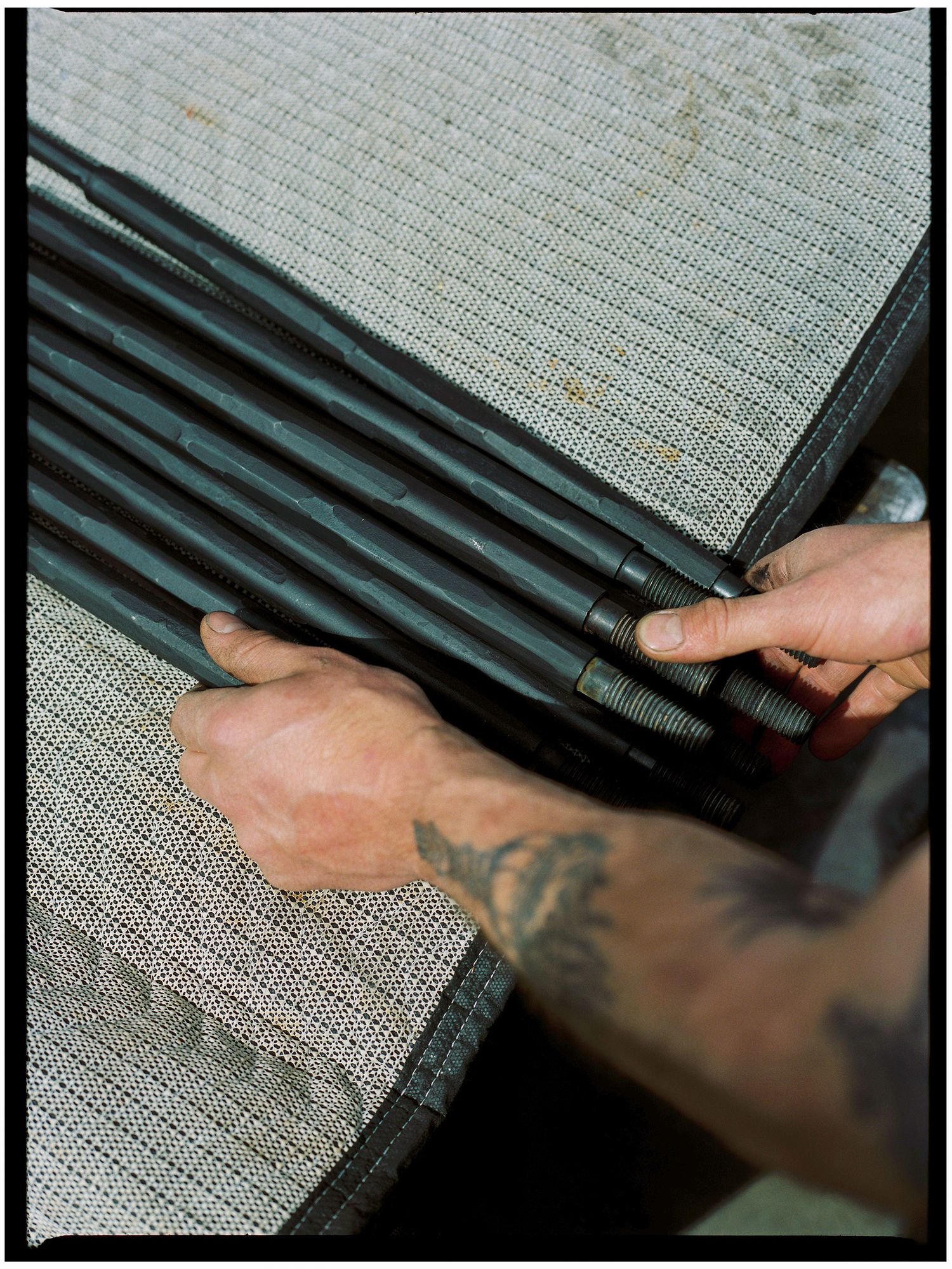 Tattooed hands hold multiple black threaded metal rods on a mesh surface.