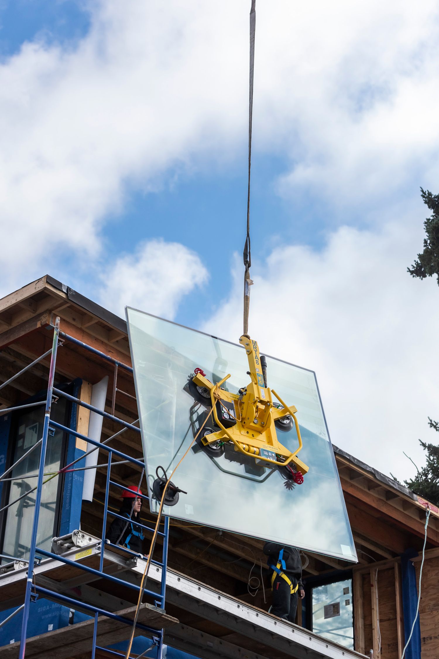 Large glass panel being lifted by yellow crane vacuum attachment during home construction.