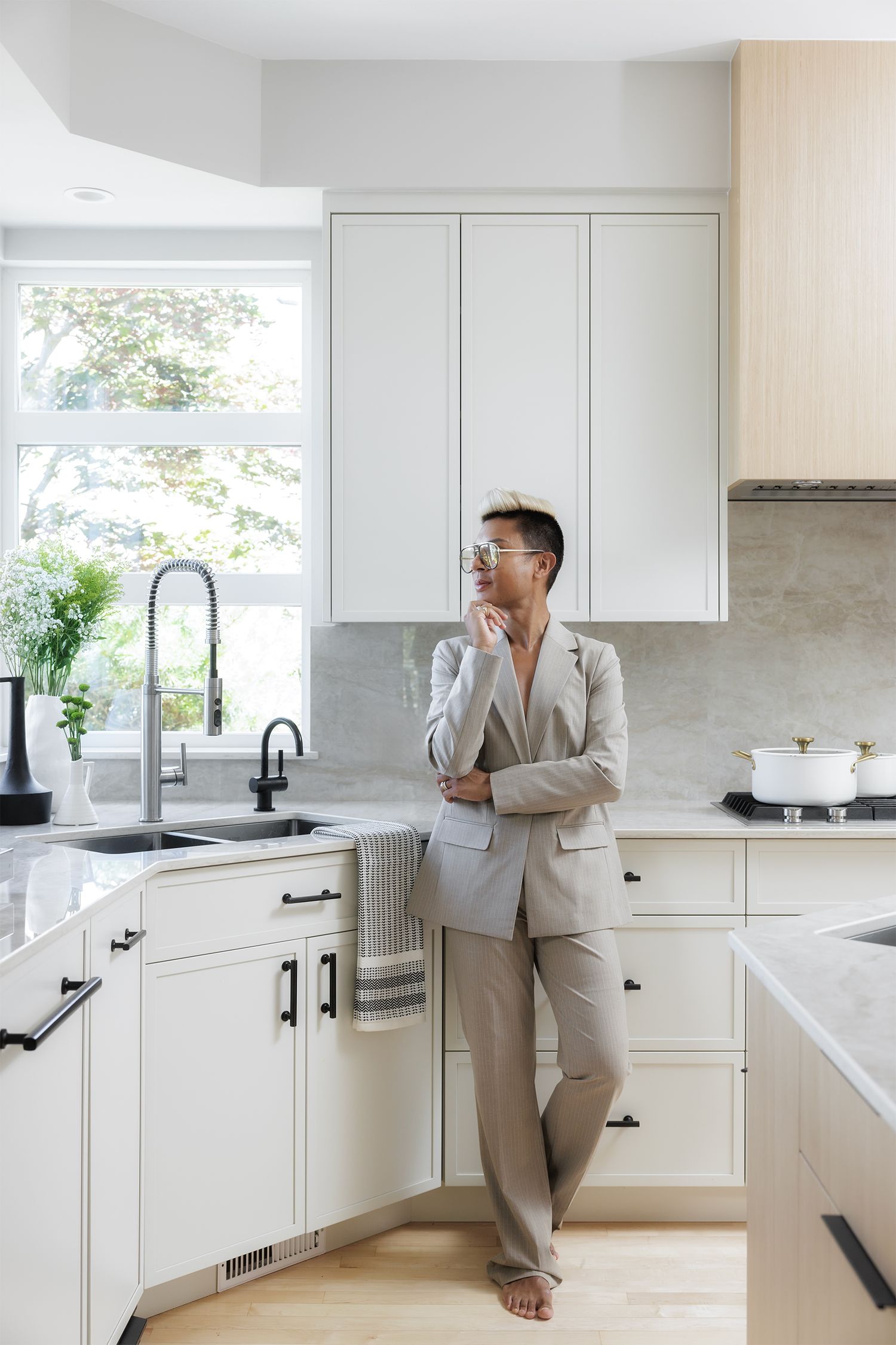 Person in beige blazer standing in bright white modern kitchen with marble backsplash and black hardware fixtures.
