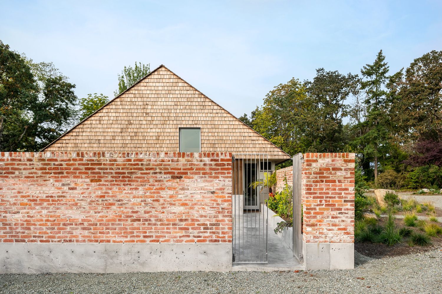 Rustic brick exterior wall with metal gate and cedar shingle roof gabled house in a garden setting.