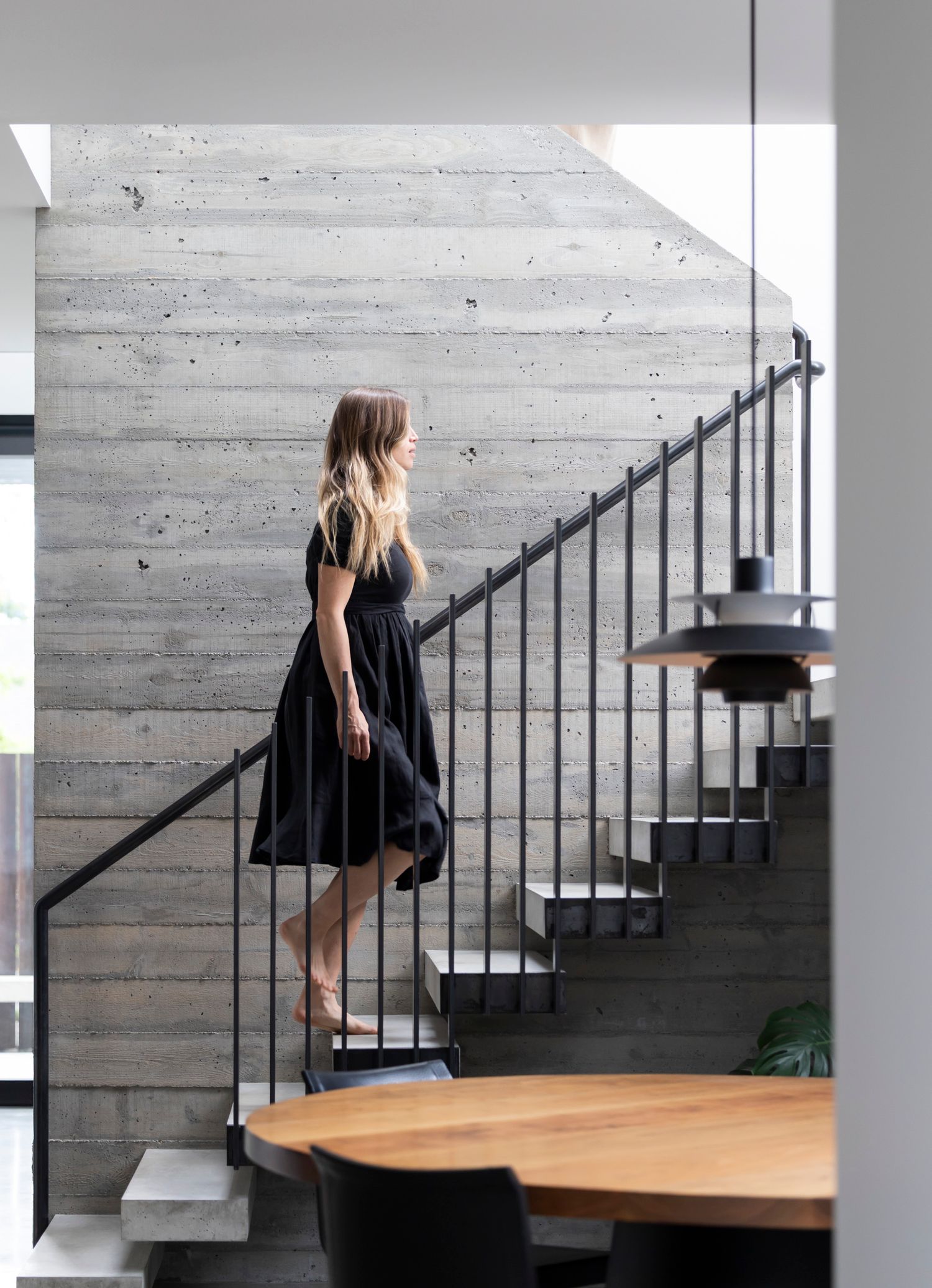 Blonde woman in black dress ascending modern concrete staircase with black metal railing beside round wooden table.