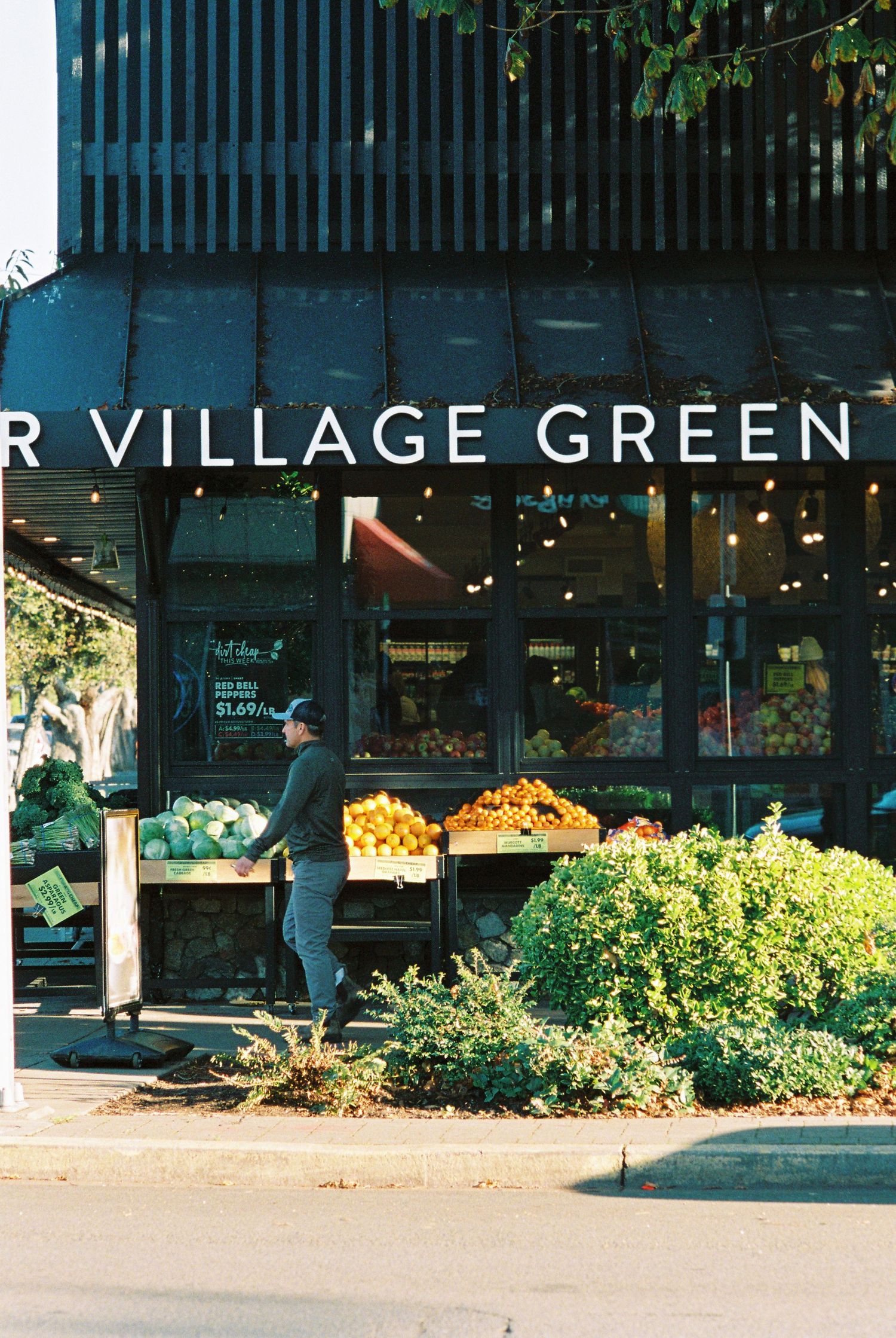 Person in black cap walks past Your Village Green grocery store with colorful outdoor produce display.