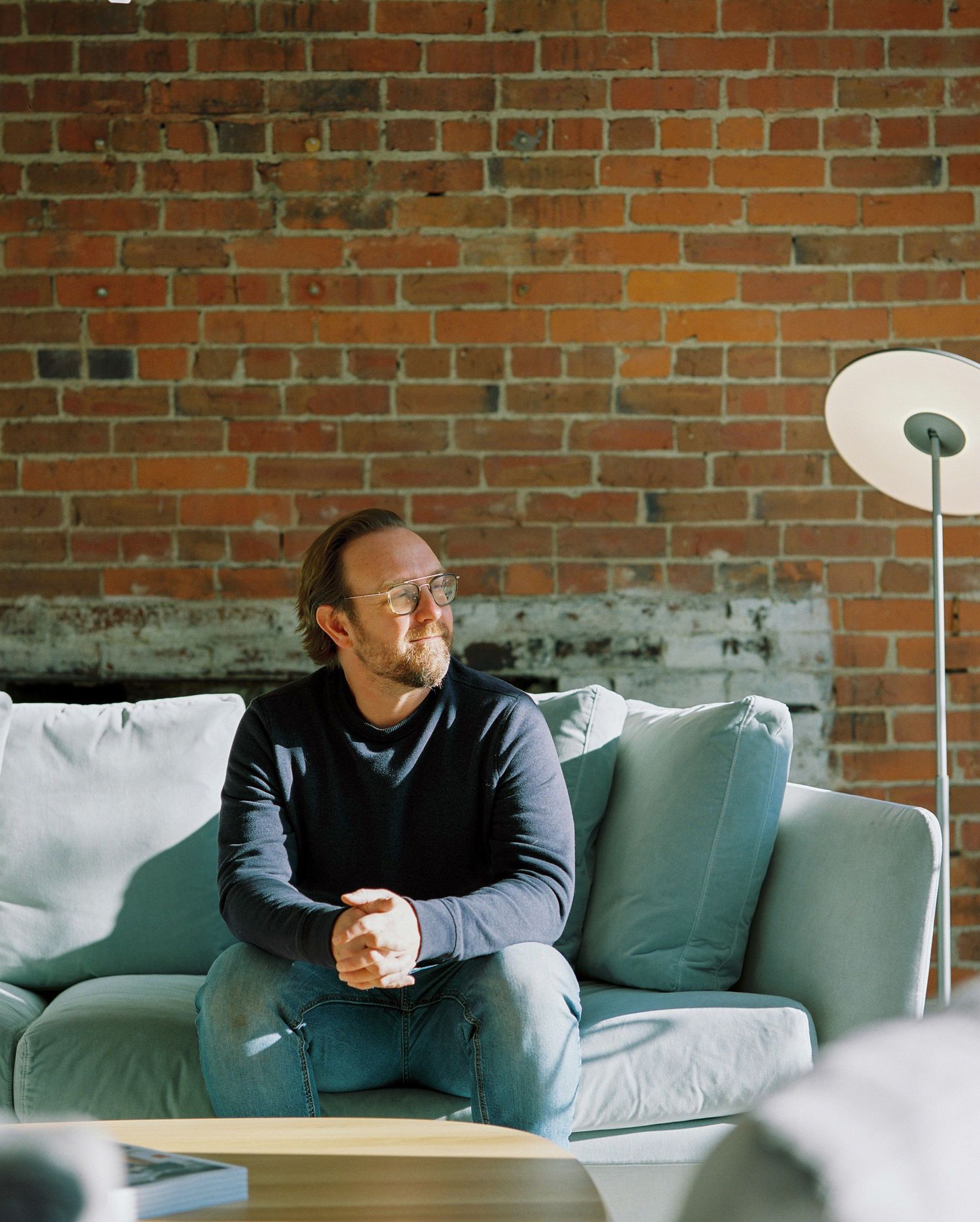 Man in black sweater and jeans sitting on grey sofa in front of exposed brick wall with floor lamp.