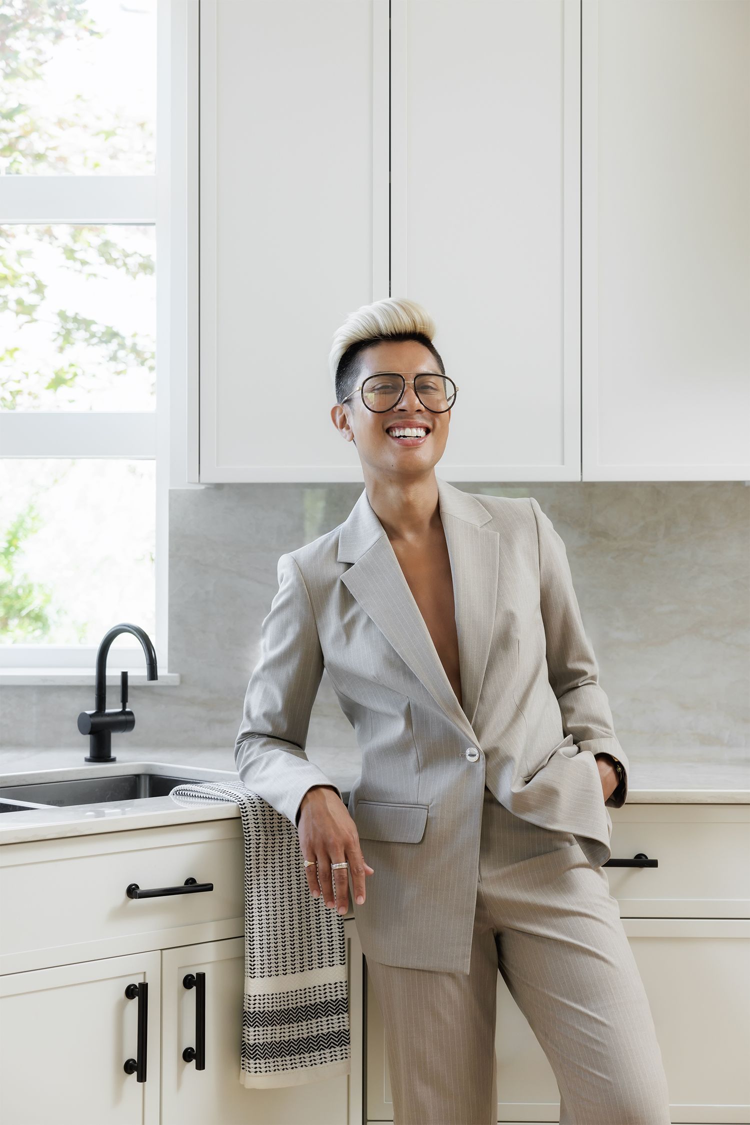 Smiling woman with short blonde hair wearing beige suit and glasses leaning against white kitchen counter.
