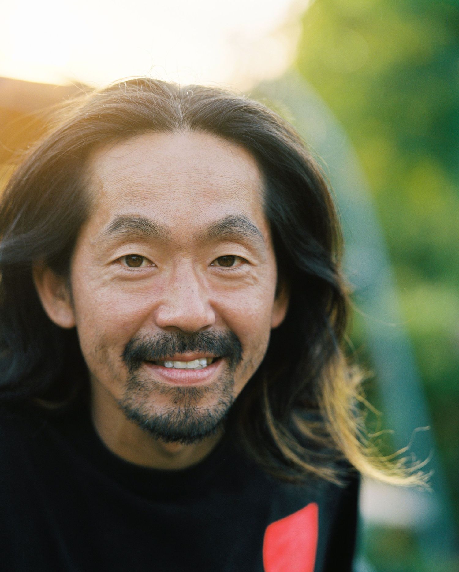 Smiling Asian man with long dark hair and beard photographed outdoors in warm golden hour light.
