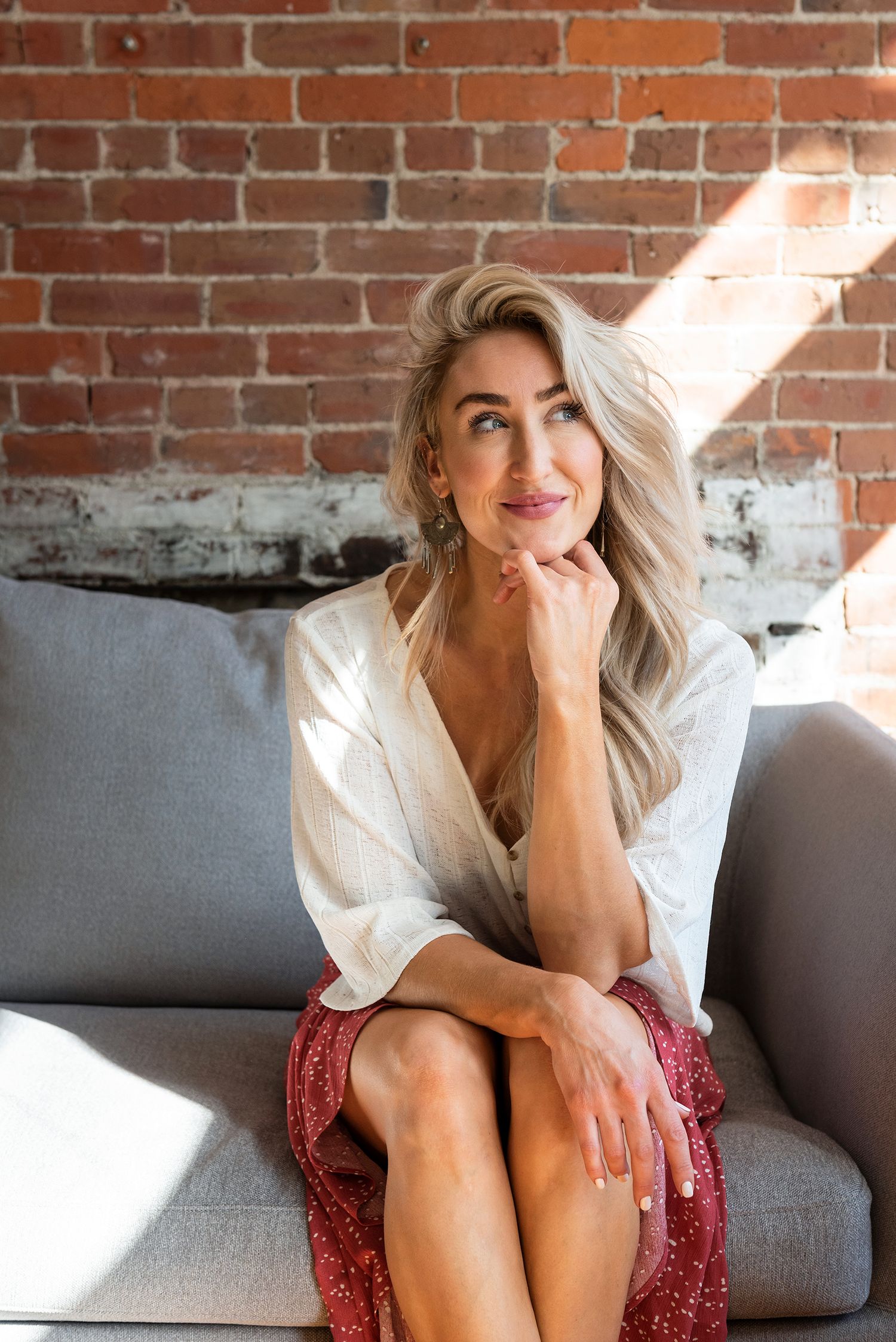 Blonde woman in white shirt and red skirt sitting thoughtfully on grey sofa against brick wall.