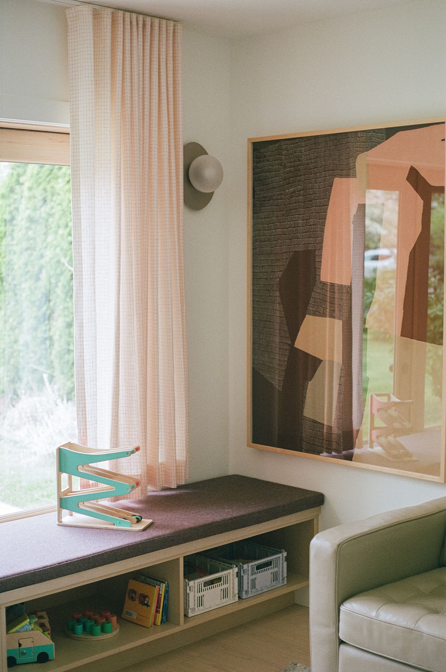 Cozy room corner with pink sheer curtains, built-in window bench with storage, abstract wall art, and natural garden views.
