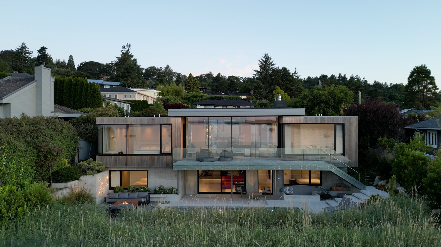 Modern two-story concrete and glass luxury home with expansive windows and balcony at dusk.