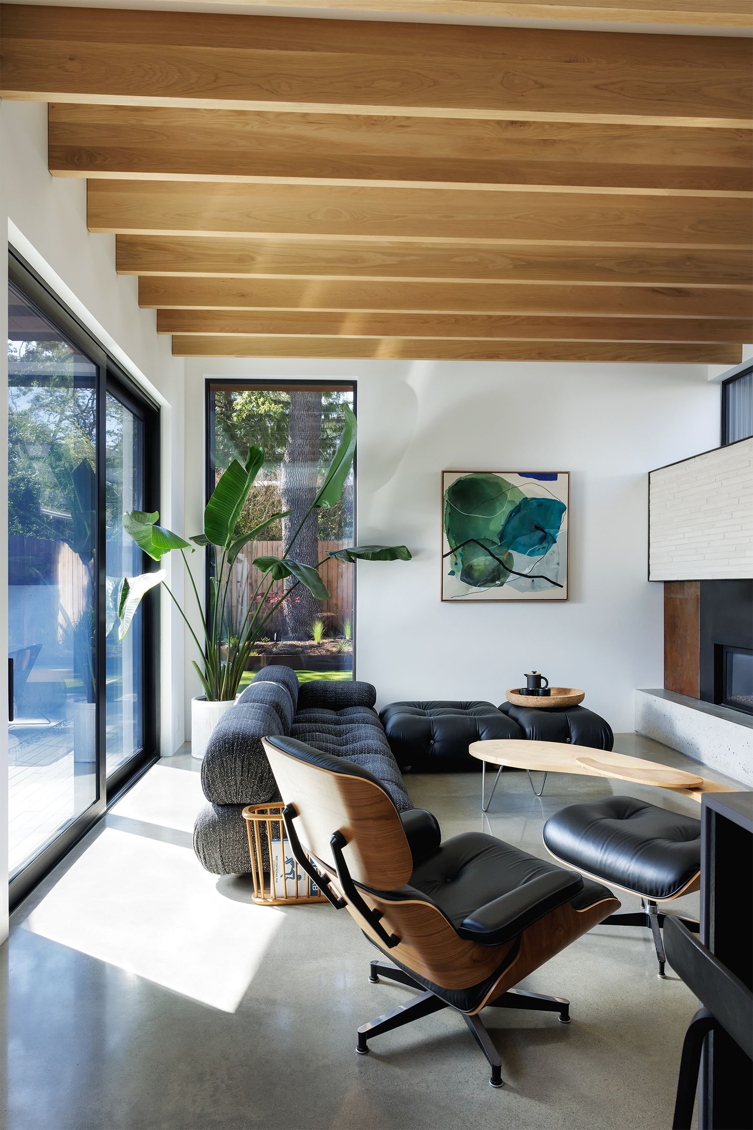 Contemporary living room with exposed wood beam ceiling, Eames lounge chair, black sofa, polished concrete floors, and tropical plant.