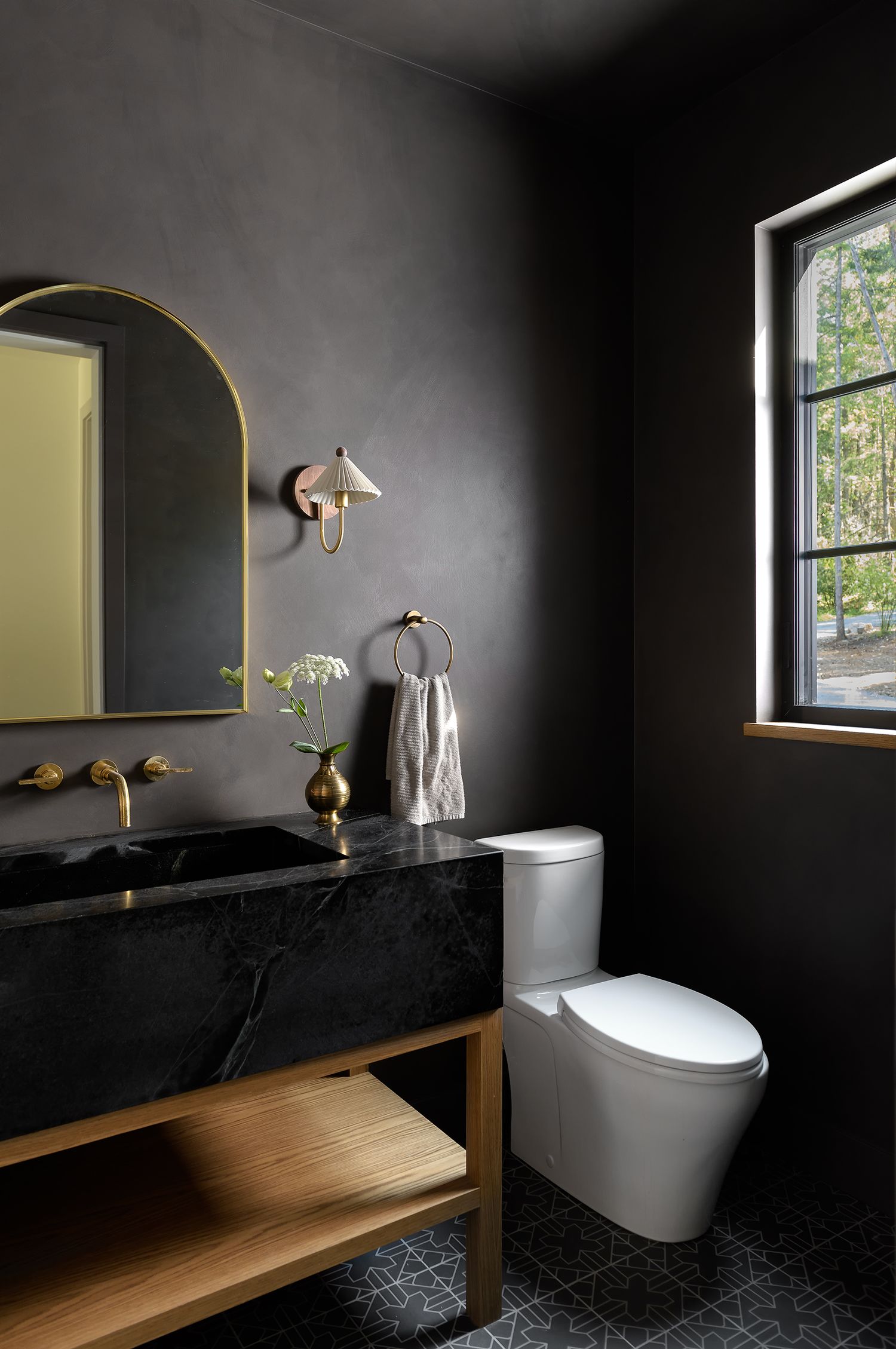 Dramatic dark bathroom with black marble vanity, brass fixtures, arched mirror, and geometric tile floor.