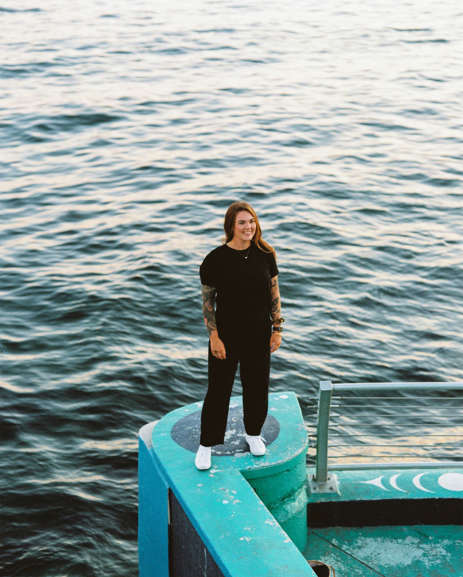 Woman in black outfit standing on turquoise dock structure with calm ocean water in background.