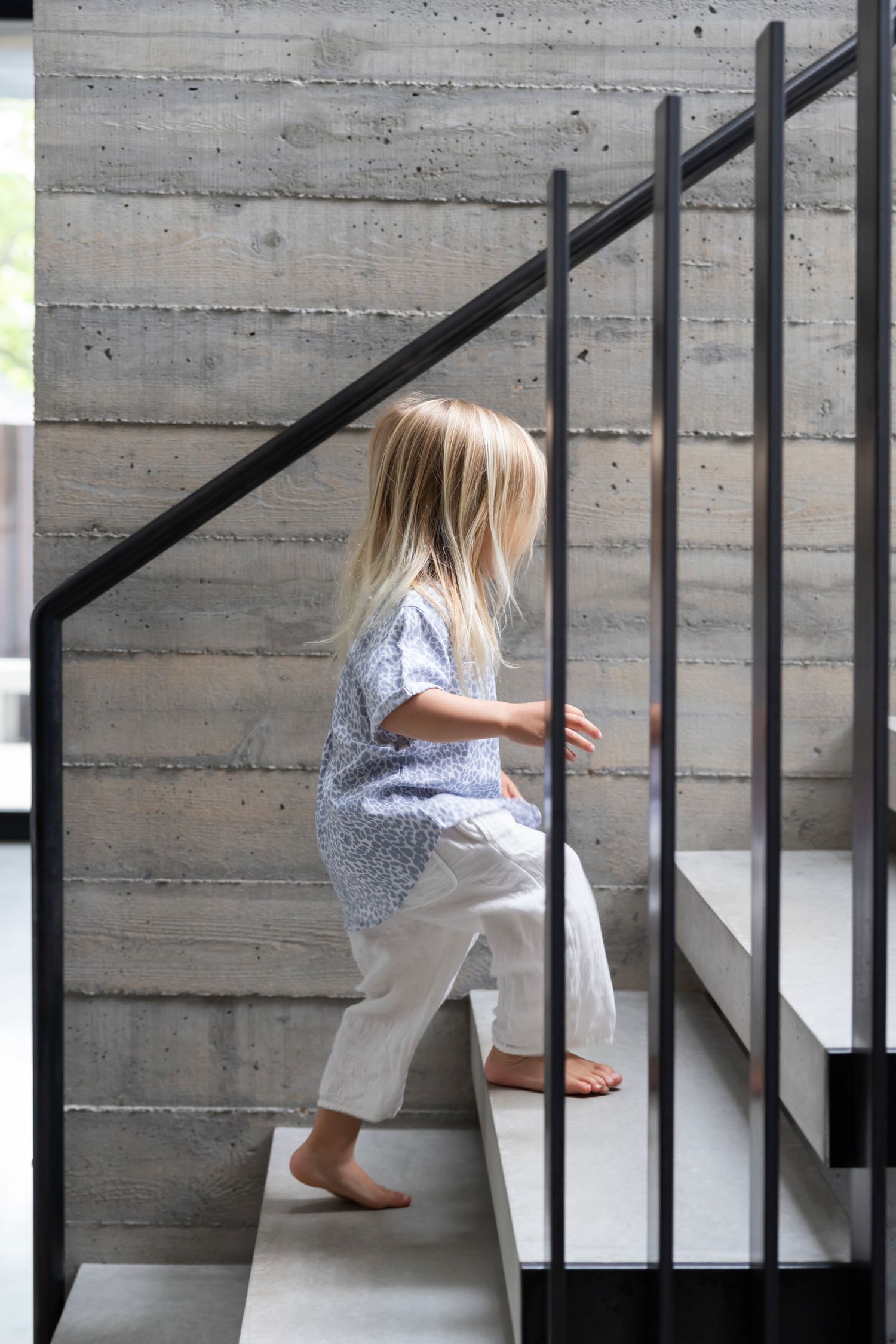 Young blonde girl in blue floral top climbing concrete stairs with black metal railing in modern home interior.