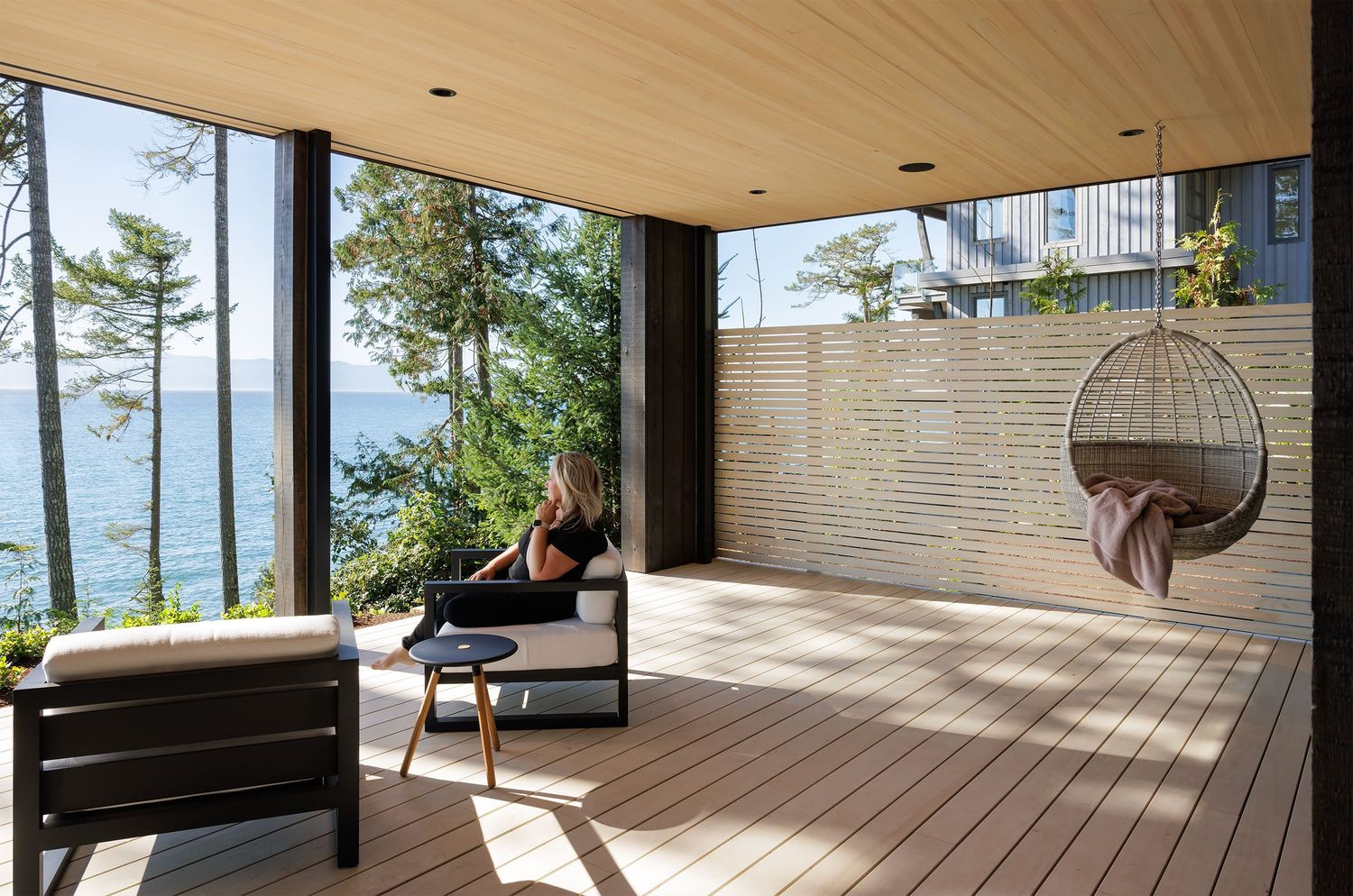 Modern outdoor deck with hanging wicker chair, wood flooring, louvered privacy wall, and panoramic water and forest views.