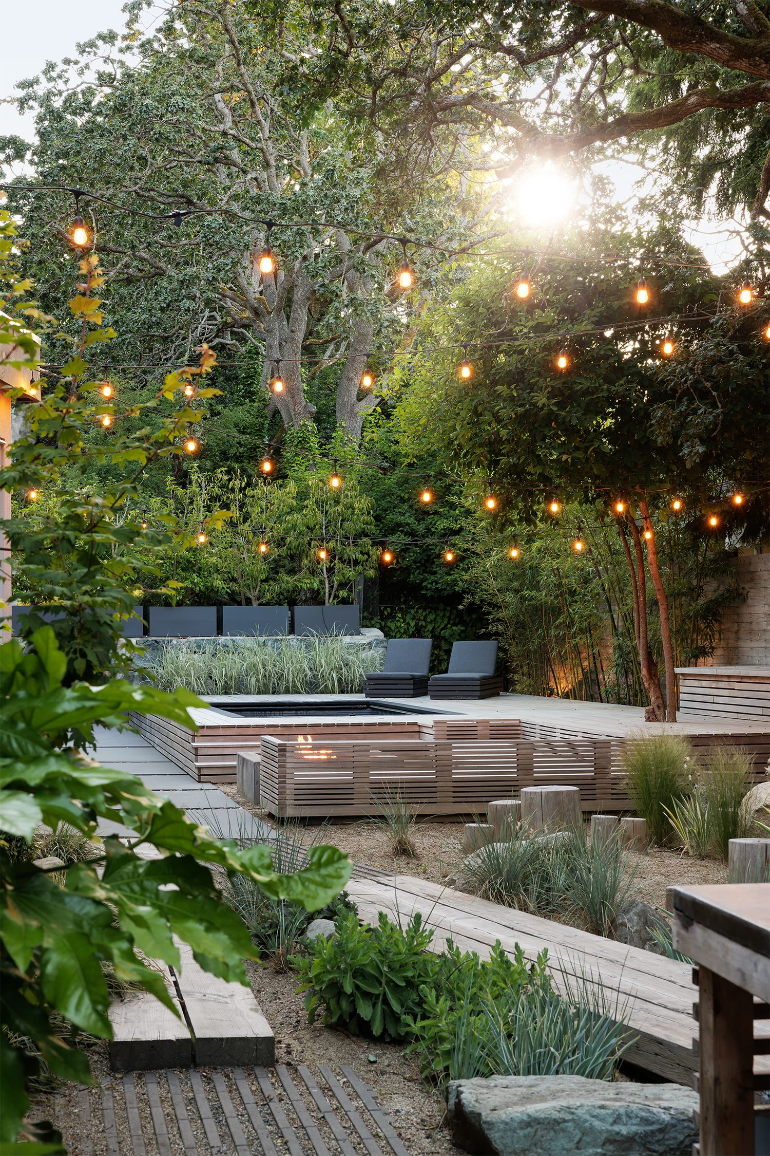 Enchanting backyard garden at dusk with string lights, tiered wooden planters, fire feature, and lush mature trees.