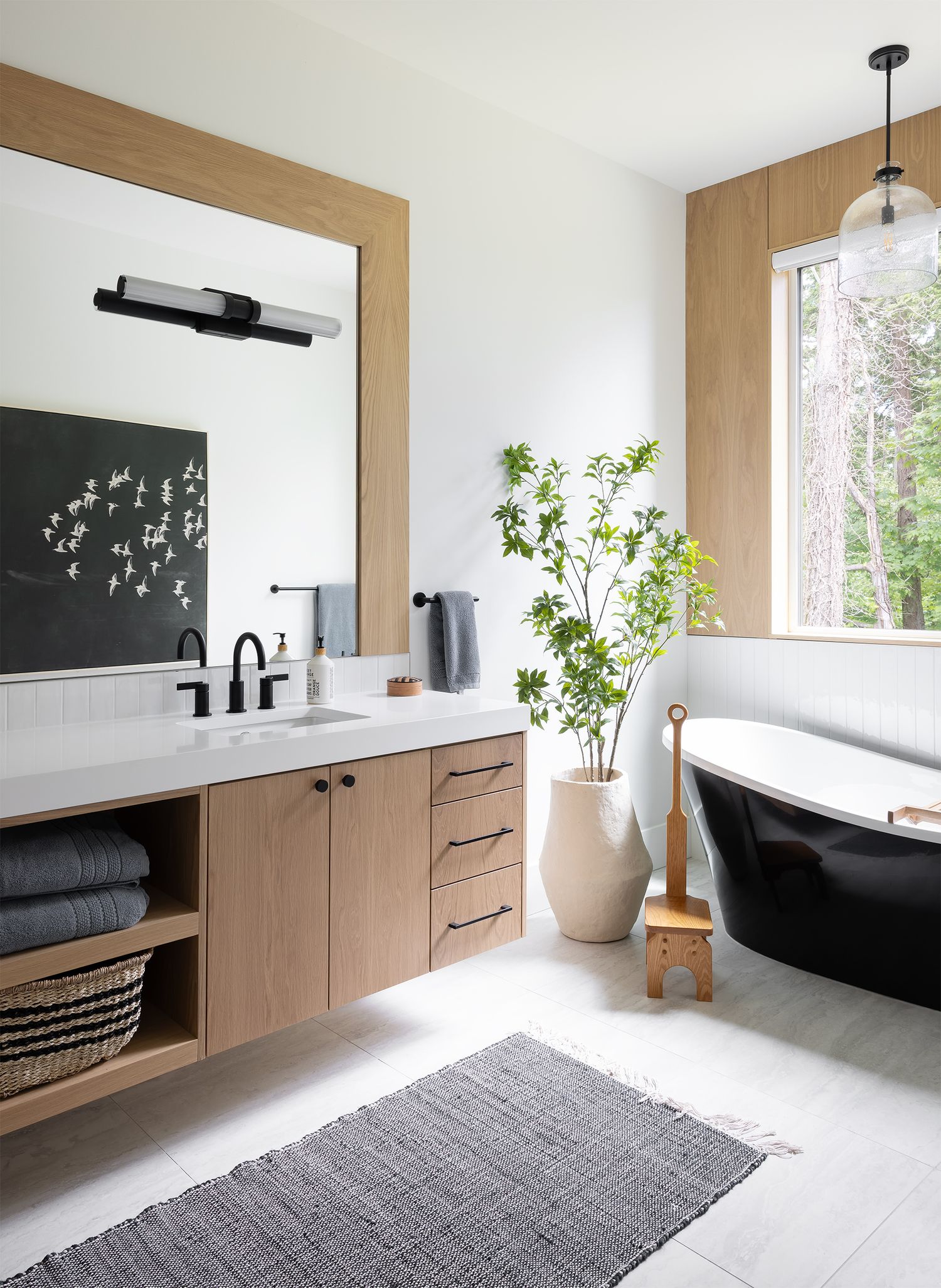 Modern bathroom with wood-framed mirror, black freestanding tub, white countertop, potted plant, and natural light.