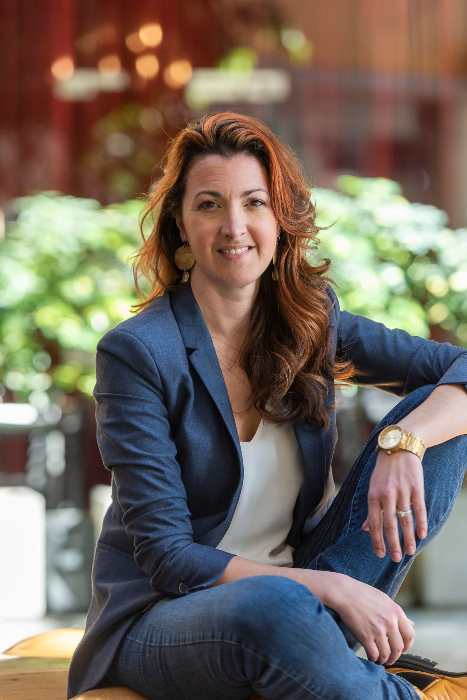 Smiling red-haired woman in blue blazer sits casually in a warmly lit indoor space.