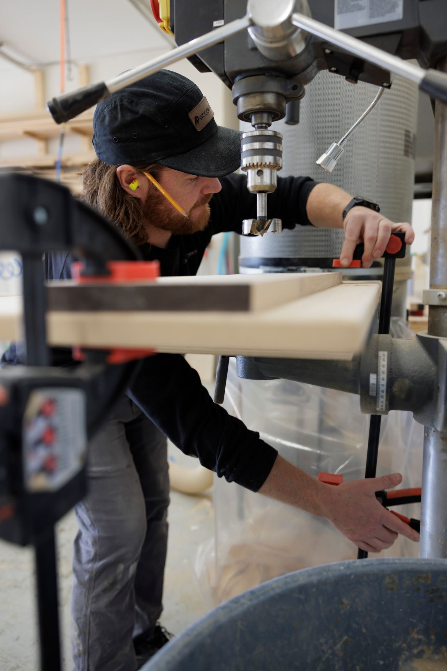 Bearded craftsman wearing safety gear operating a drill press on wood plank in a woodworking workshop.