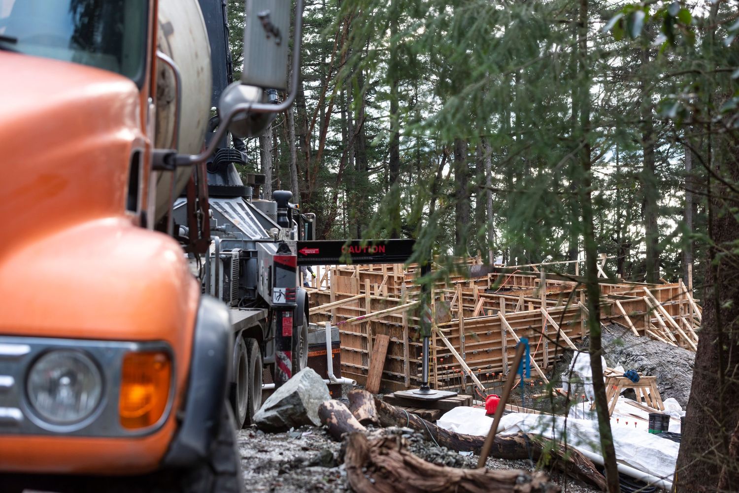 Orange cement mixer truck parked beside wooden concrete formwork structure in a forested construction site.