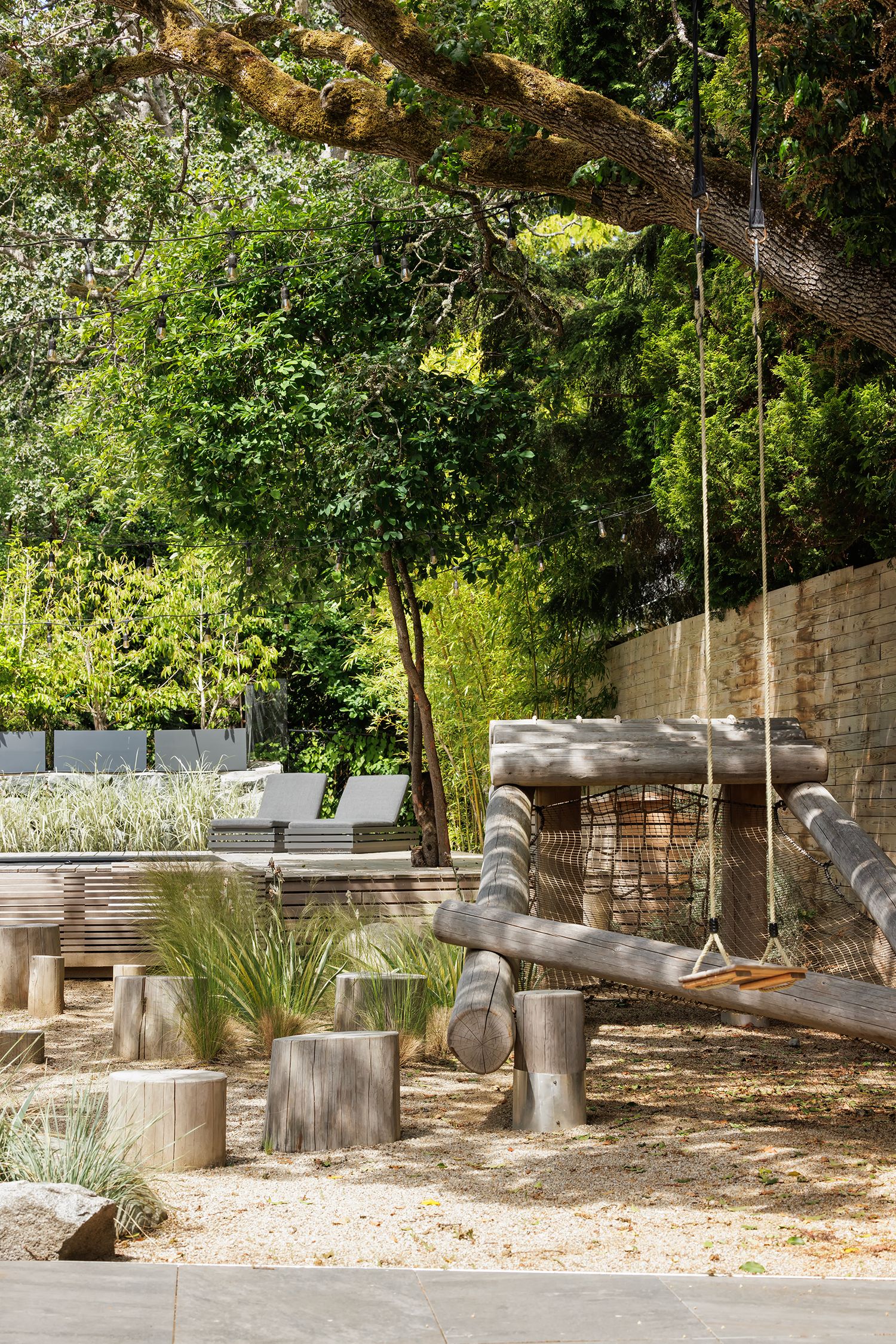 Natural outdoor playground with wooden log stumps, rope swing, and lush green trees in a landscaped garden setting.