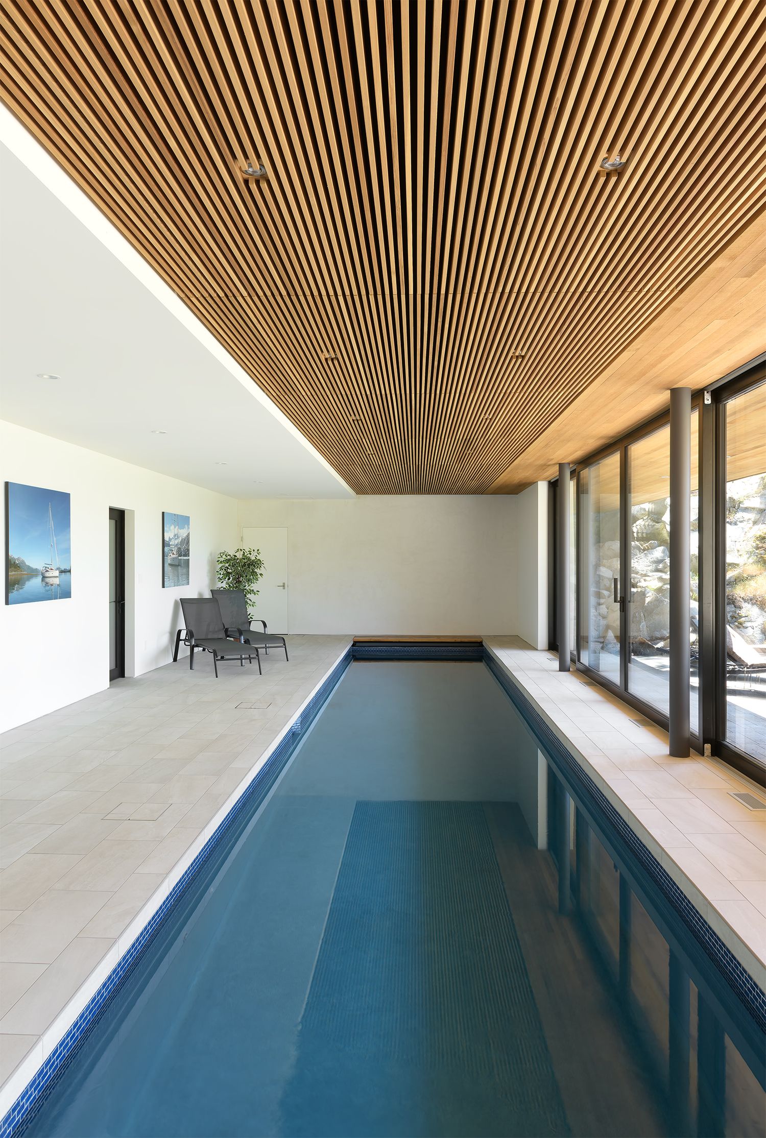 Elegant indoor lap pool with wooden slat ceiling, large glass doors, and minimalist poolside seating in a luxury home.