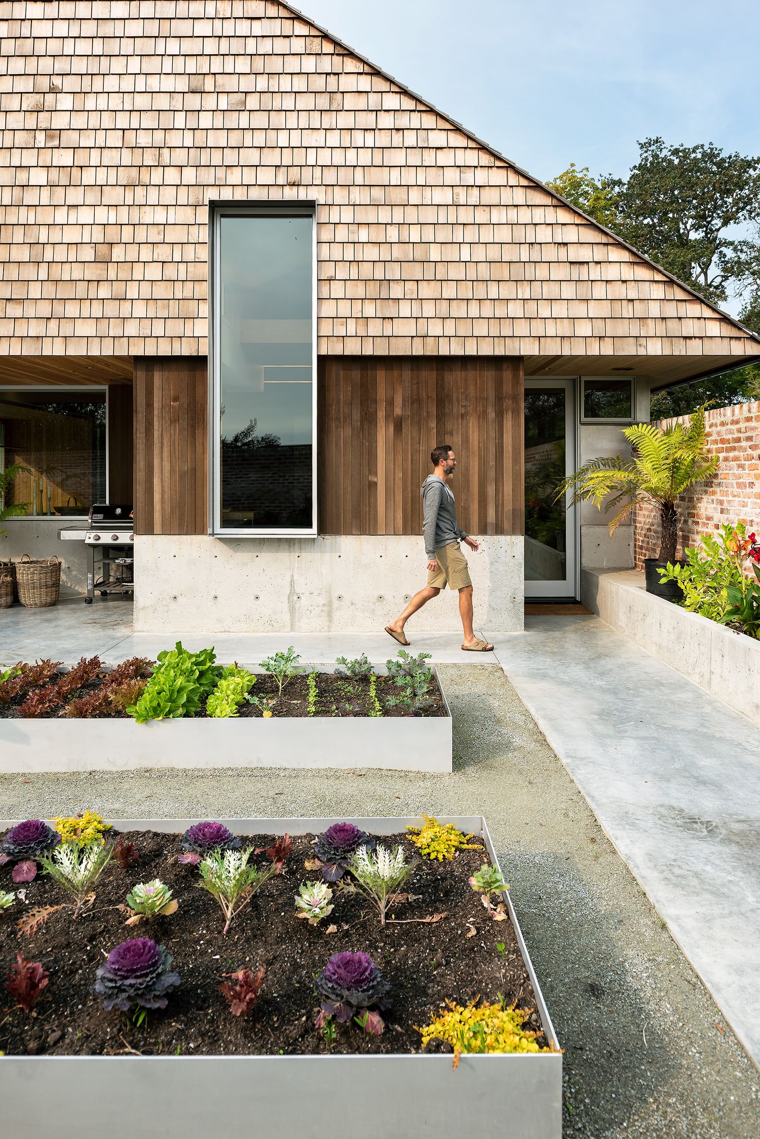 Person walking past a cedar shingle home with raised concrete garden beds filled with colorful plants.