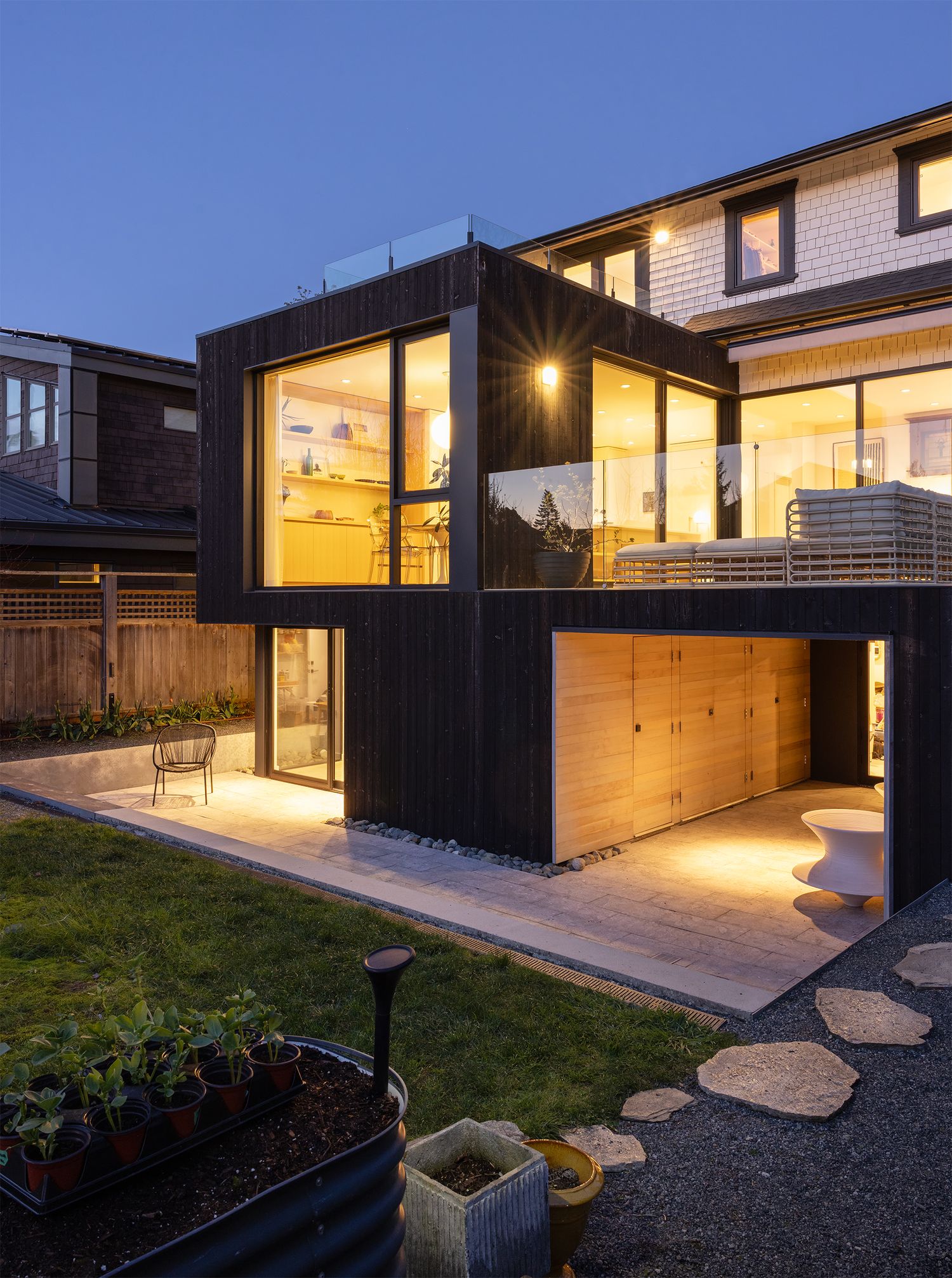 Modern home exterior at dusk with dark wood cladding, large illuminated windows, and landscaped backyard.