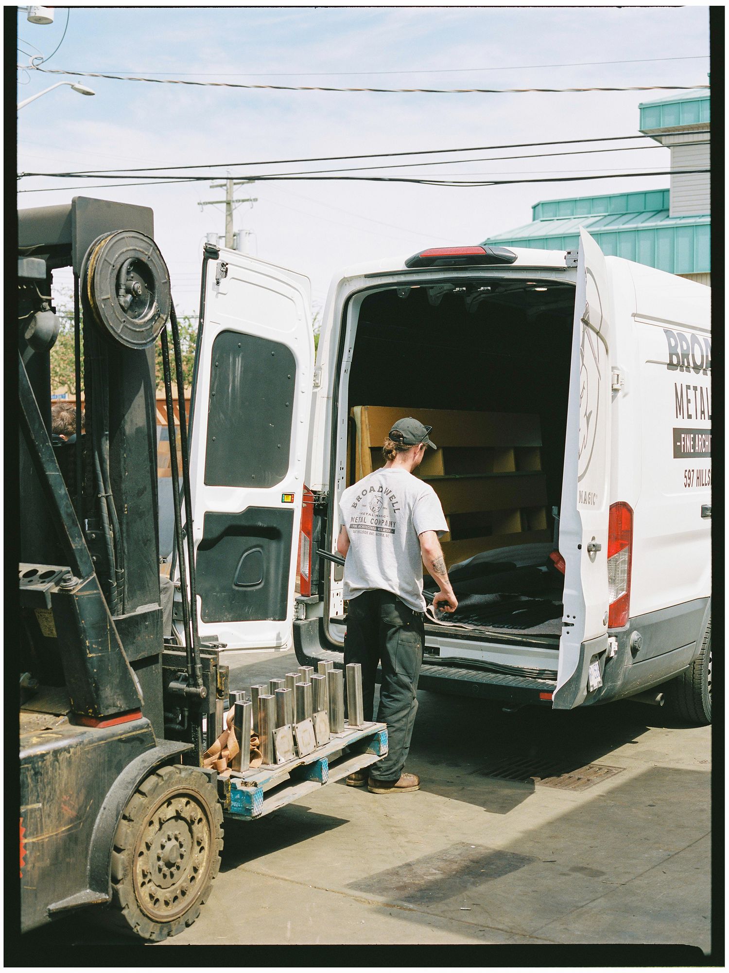 Worker loads cargo into a white Broadwell Metal Company van beside a forklift.